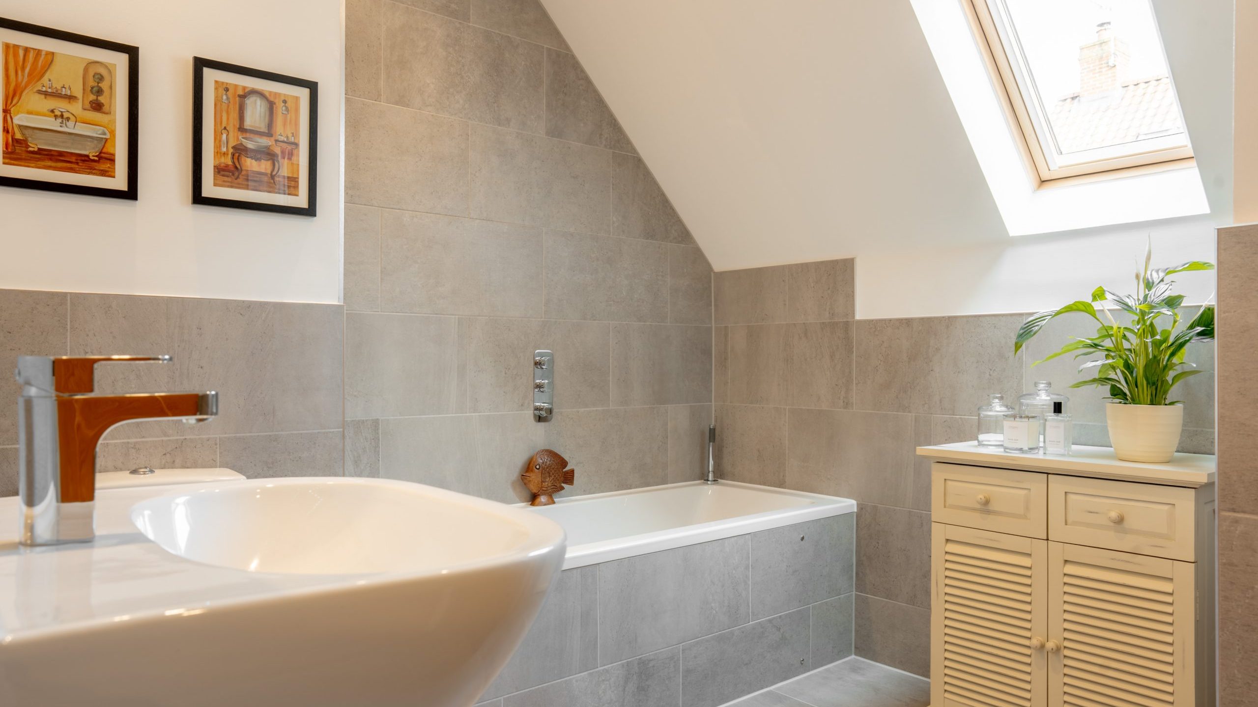 A modern bathroom with a slanted ceiling features a bathtub, a white vanity with a plant, and a skylight. Two framed artworks hang above the tub, and the walls and floor are tiled in gray. A white basin is in the foreground.
