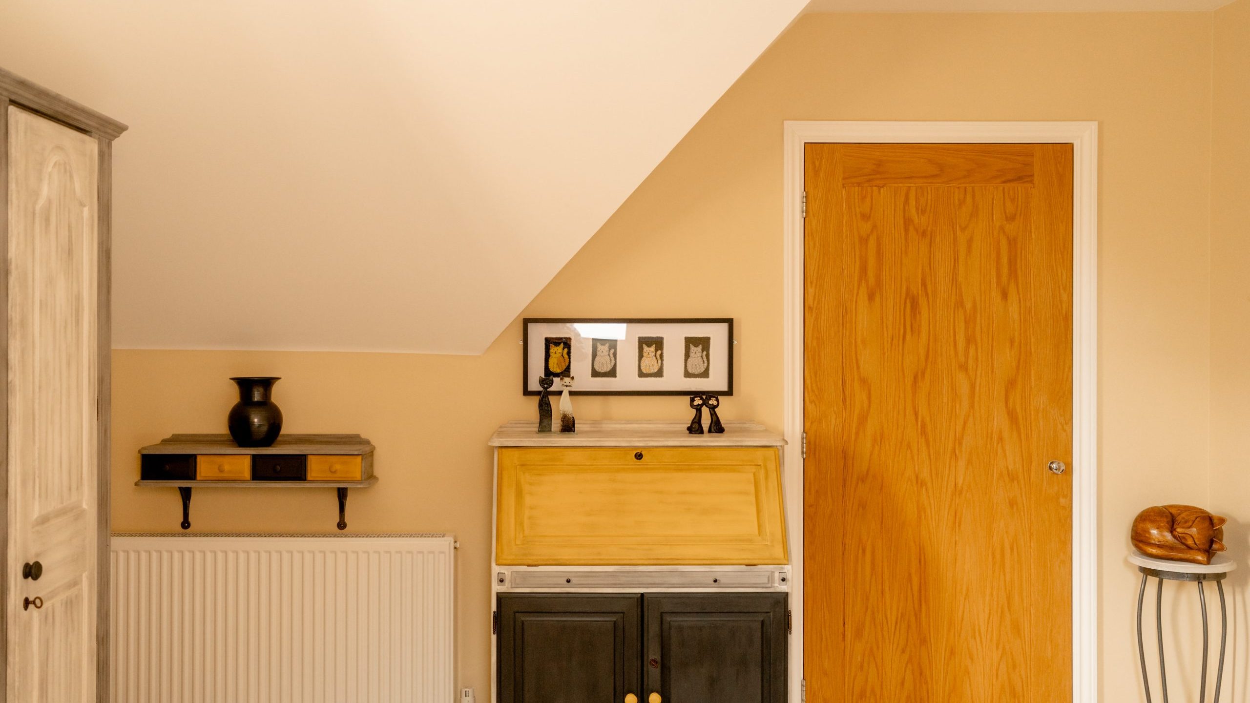 A cozy room with slanted ceiling features a wooden cabinet and a yellow bureau below framed photos. A wooden door is adjacent, and a small black table holds a sculpture. A radiator is under a shelf with decorative items. Walls are painted beige.