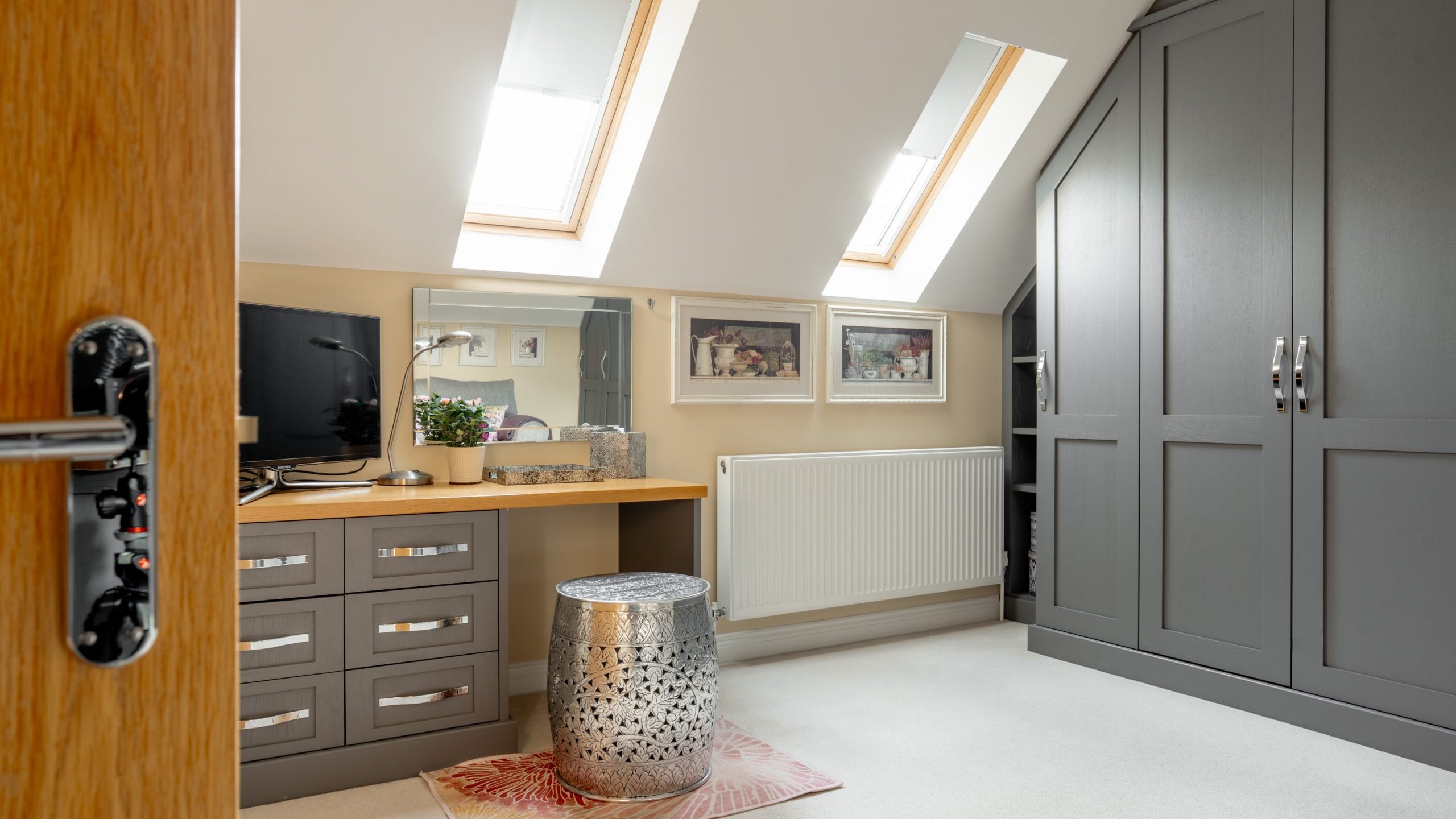A cozy attic room with skylights, featuring a large gray wardrobe and a wooden dressing table with a mirror and a small TV. A metallic stool sits on a patterned rug, and framed photos hang on the cream walls.