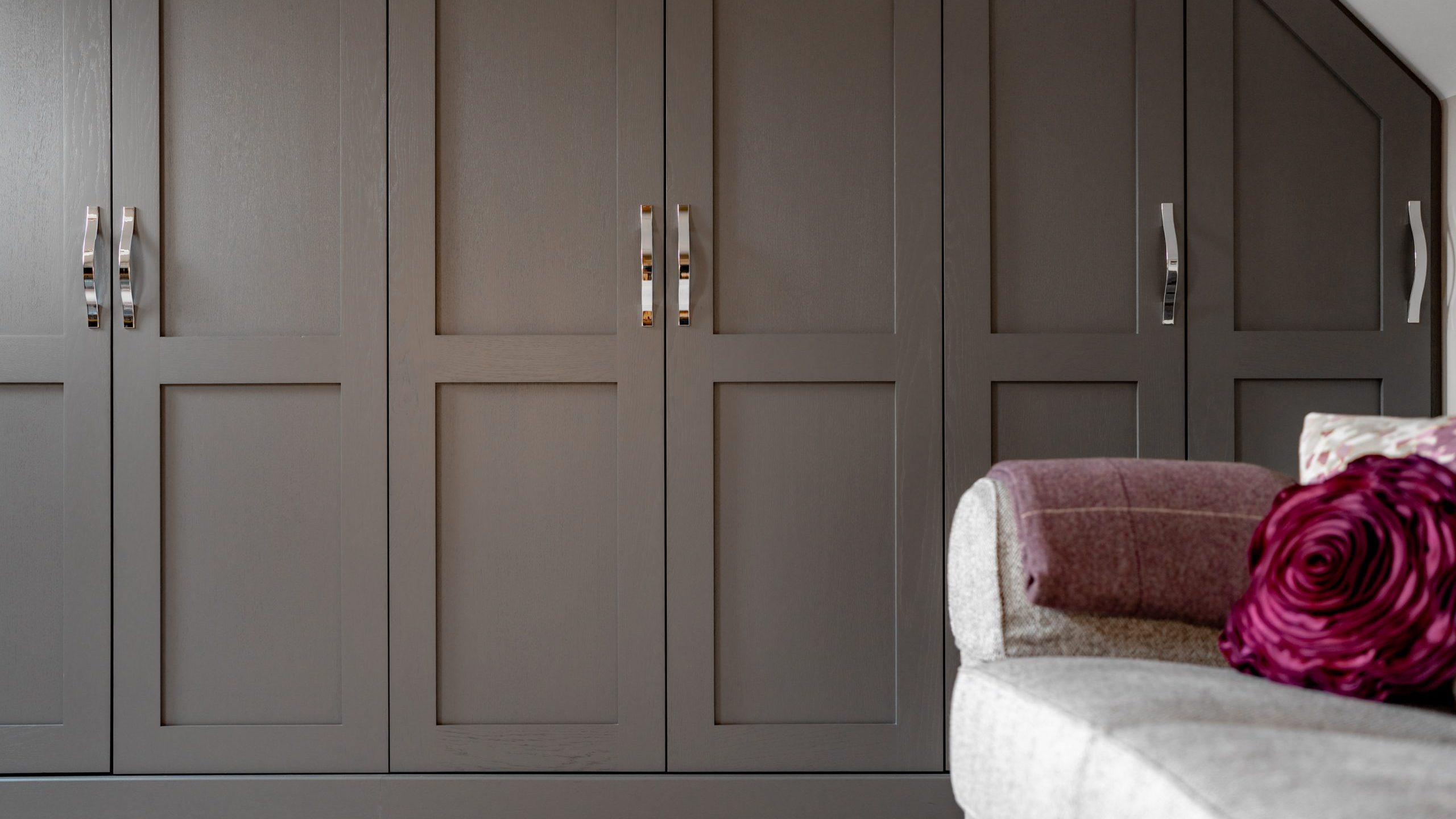 Built-in wooden wardrobe with dark brown doors and silver handles spans the wall. A light gray sofa is partially visible in the foreground, adorned with a purple textured pillow and a folded purple blanket. The room has a modern, cozy feel.