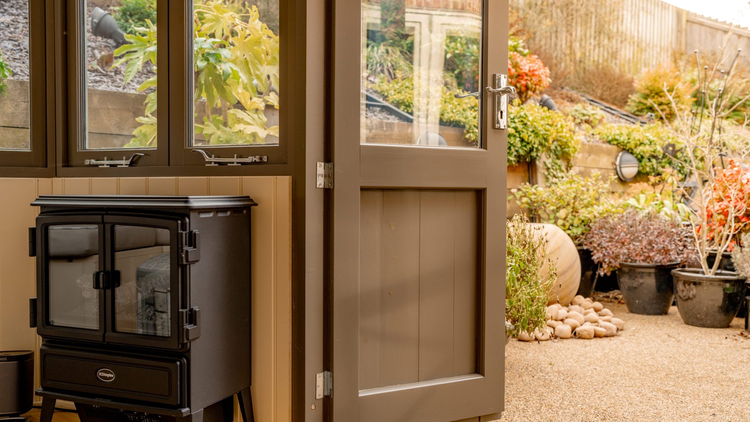 A cozy room with a black wood stove next to partially open brown double doors, leading to a garden filled with various plants and shrubs. A stone path and a large potted plant are visible outside under a wooden fence.