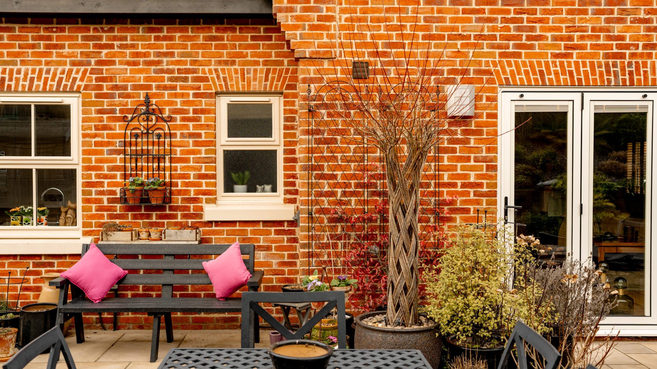 Brick house patio with a black bench adorned with two pink pillows. The scene includes potted plants, a black metal table and chairs, and glass double doors. Nearby, decorative wire wall art and a small window with plants are visible.