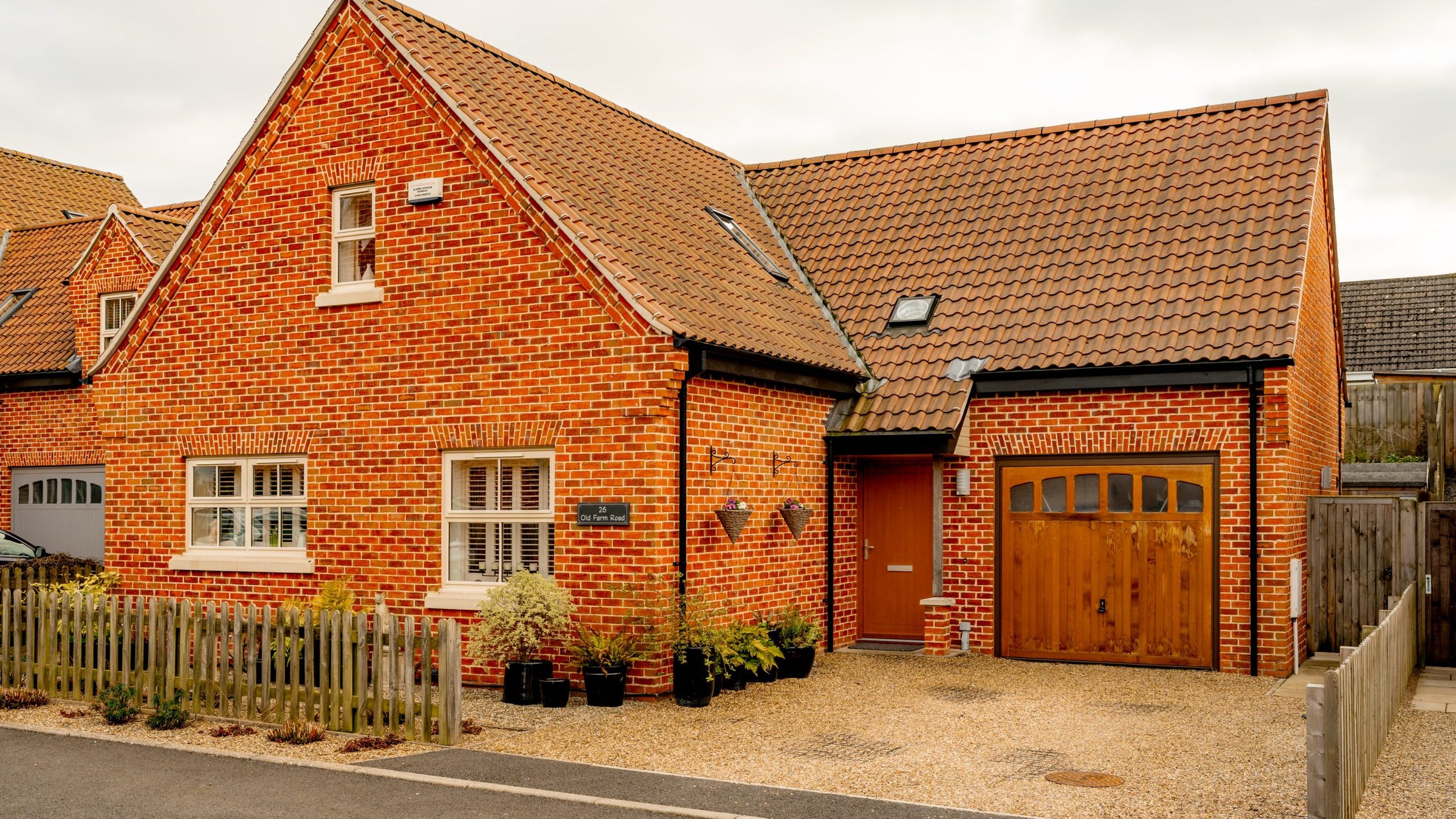 A red brick house with a brown tiled roof and attached garage. There's a wooden fence and several potted plants in front. The garage door is wooden, and the main entrance is to the right of the house. The sky is overcast.
