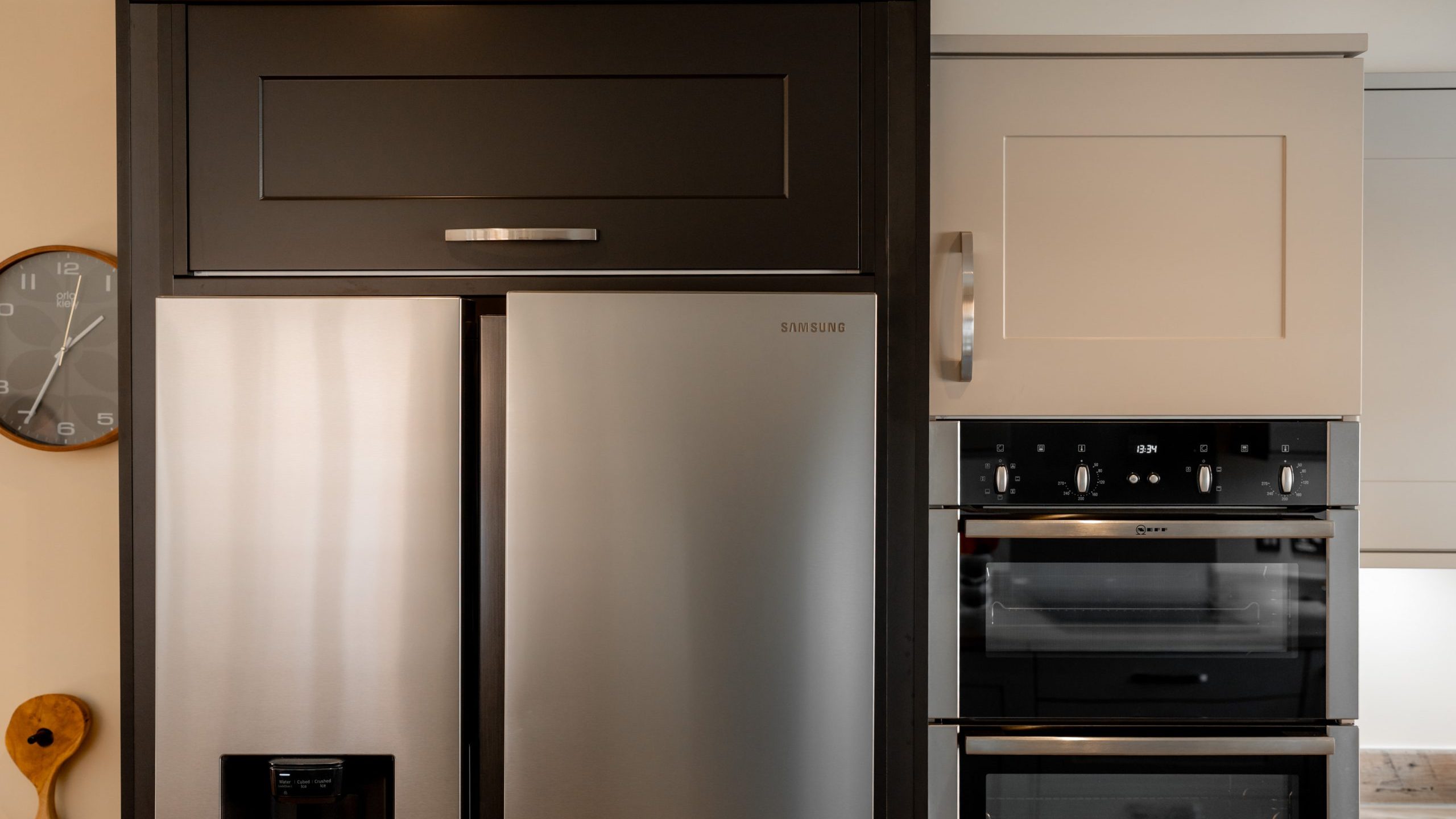 A modern kitchen features a stainless steel refrigerator and a built-in oven next to it. A wall clock is partially visible on the left along with a wooden cutting board with a handle. The cabinets are dark and light gray.