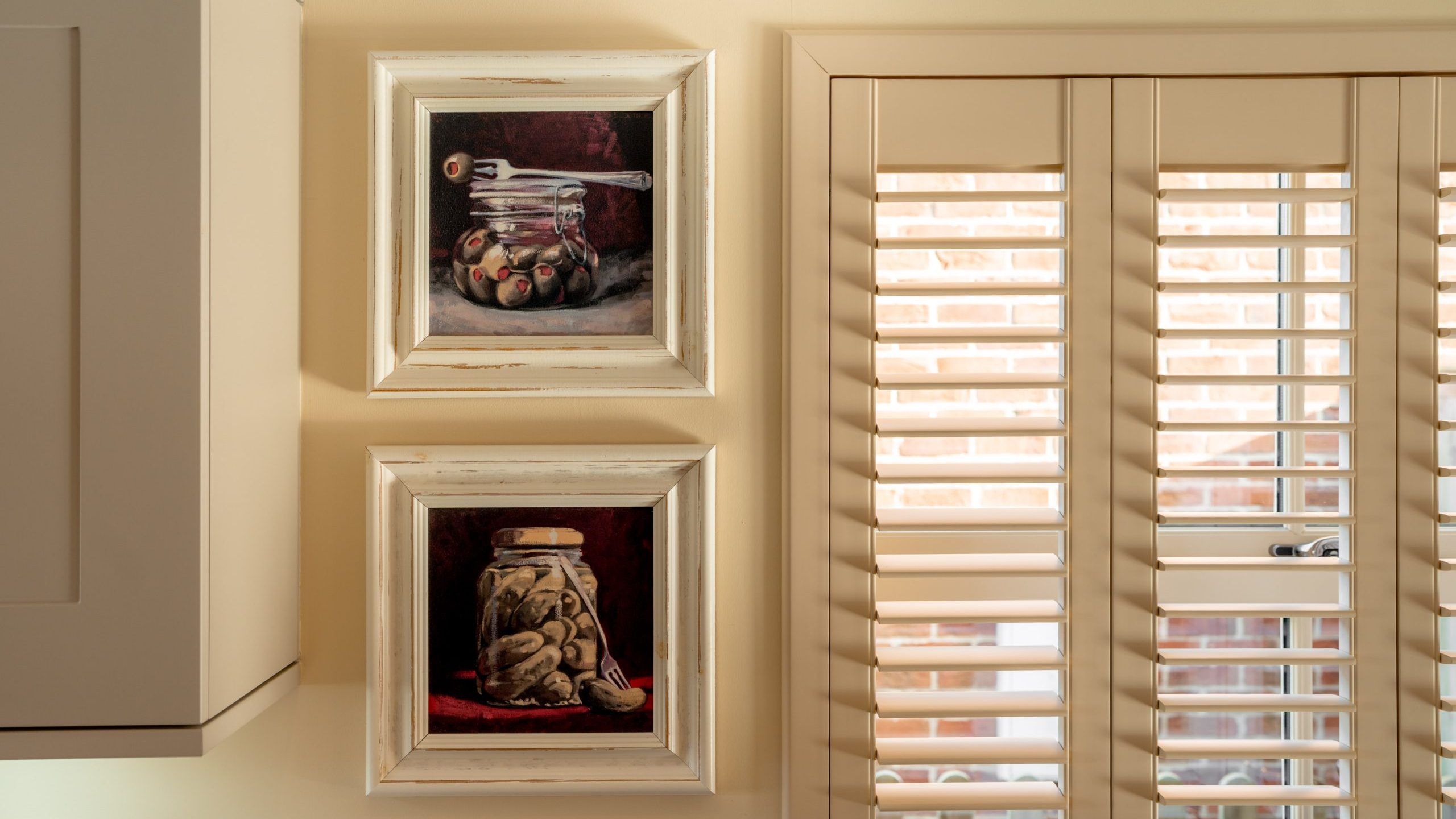 Two framed paintings of jars with spoons and fruit hang on a cream-colored wall next to white plantation shutters with sunlight filtering through. A section of a cabinet is visible on the left.