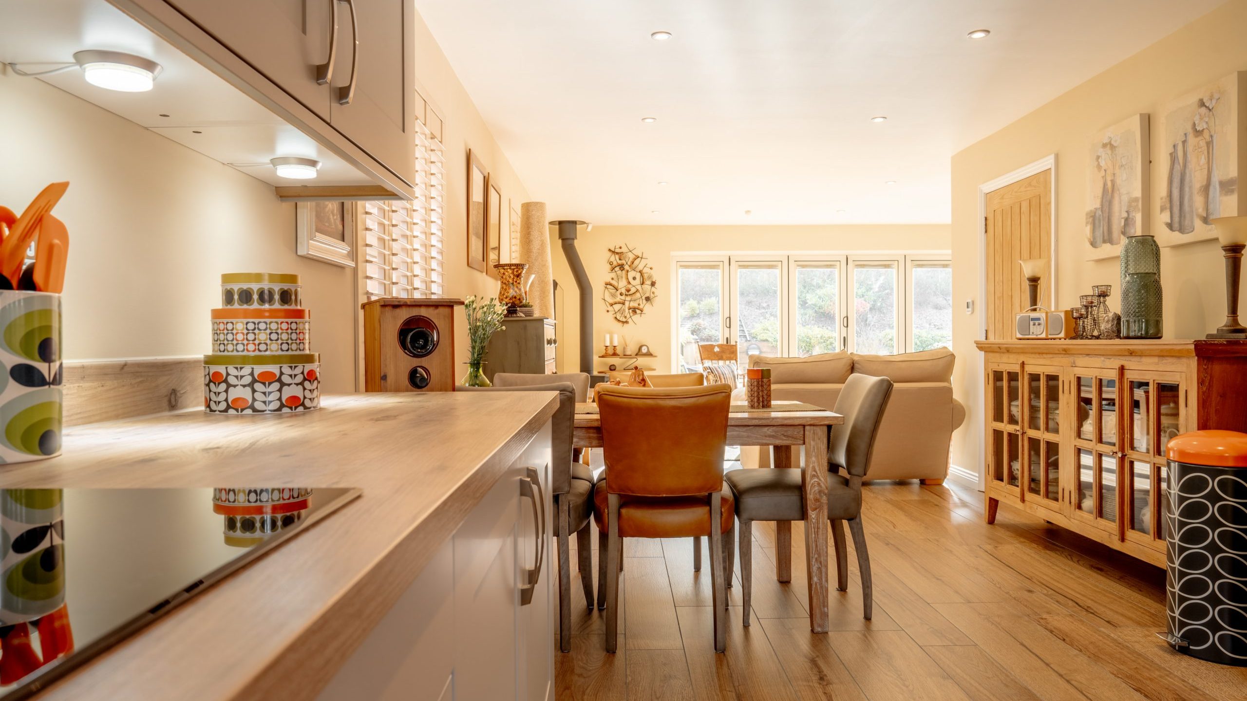 Modern open-plan kitchen and dining area with wooden flooring. The kitchen features a sleek countertop and patterned containers. The dining space has a wooden table with leather chairs, leading to a cozy sitting area and a large window with garden views.