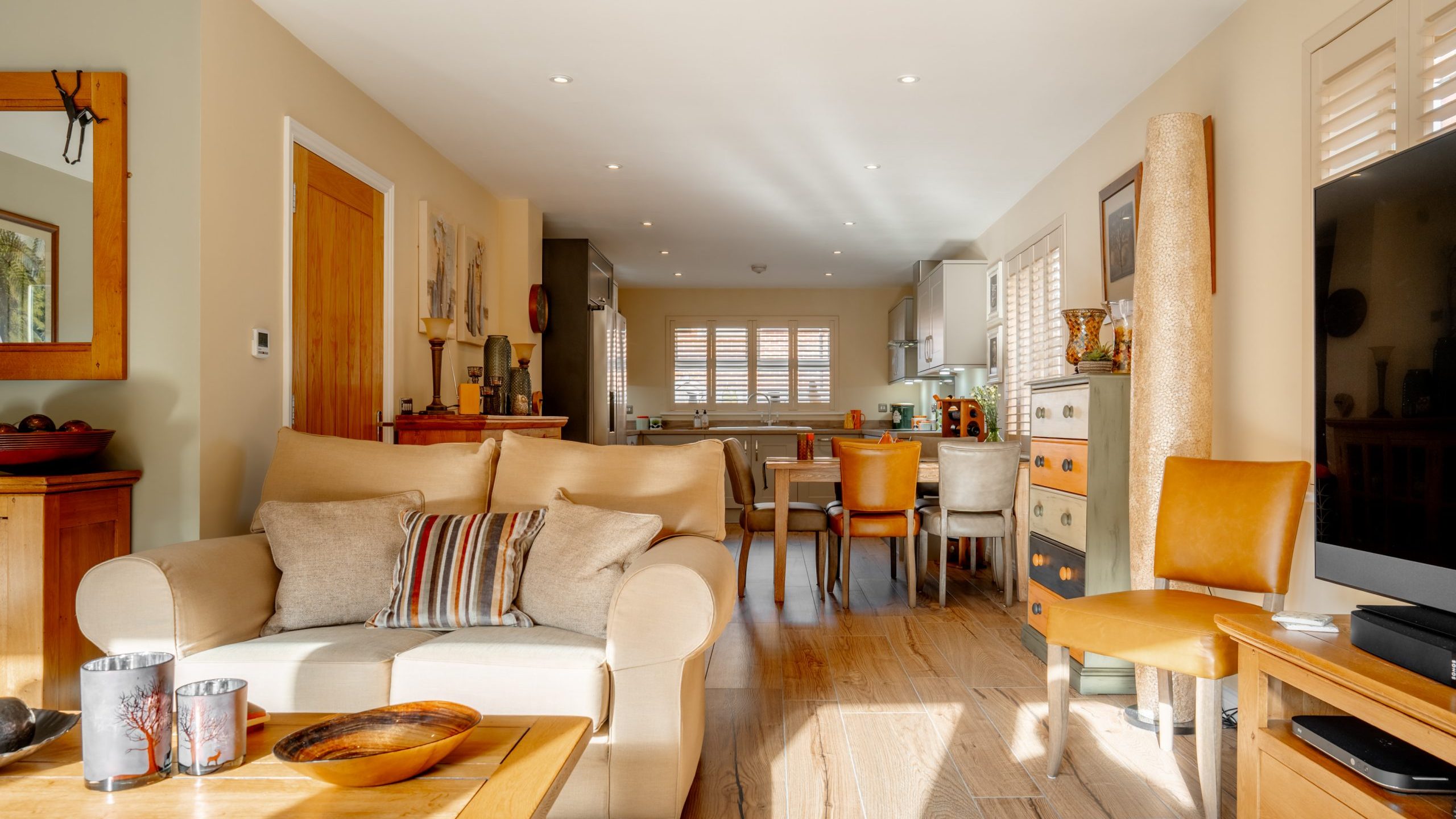 A cozy living room and kitchen area with beige walls and wooden flooring. It features a beige sofa with striped cushions, a wooden coffee table, various chairs, a cabinet, and a large flat-screen TV. Sunlight streams in, brightening the space.