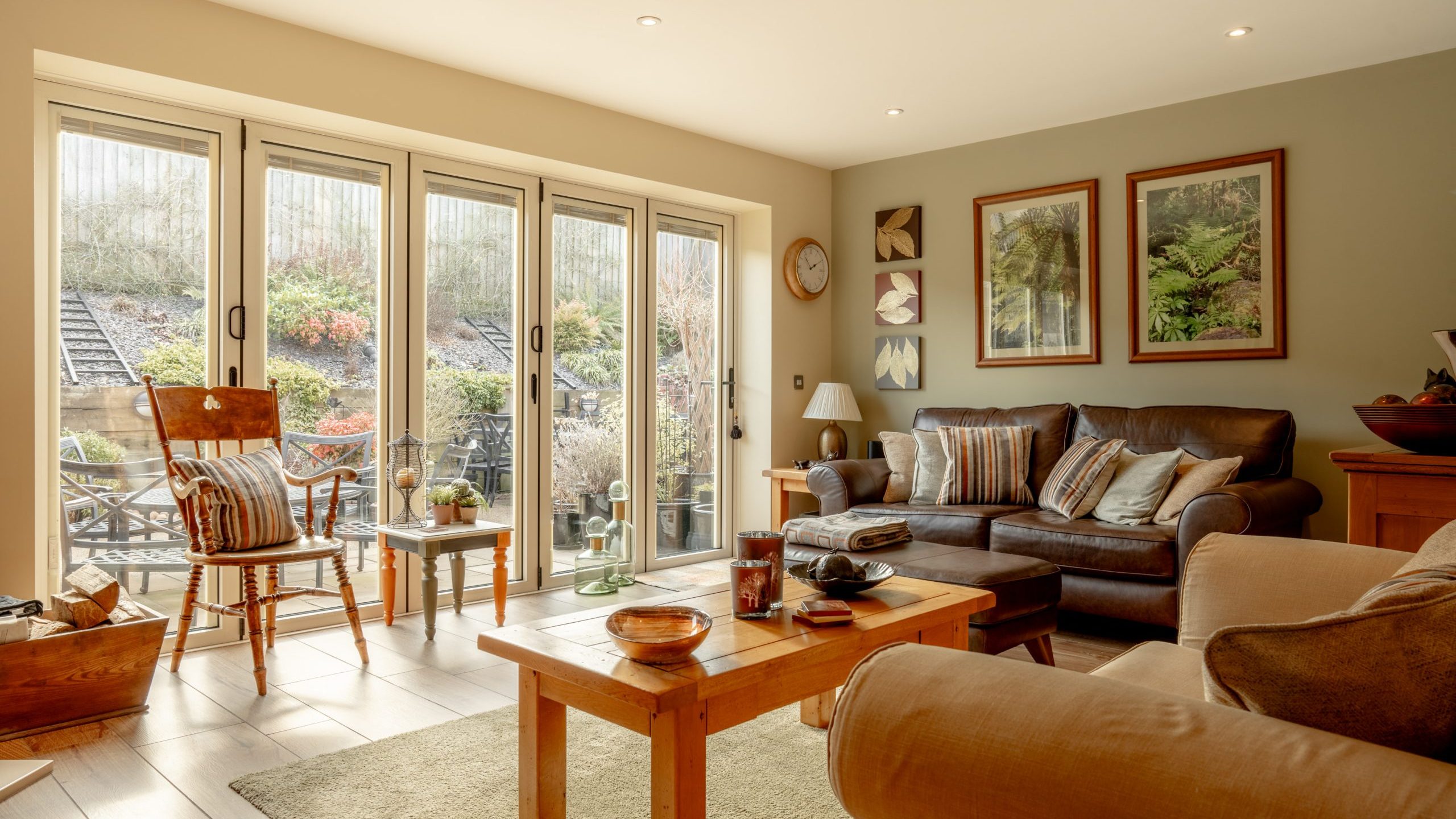 A cozy living room featuring large glass doors opening to a patio. It has comfortable seating with a sofa, armchair, and a rocking chair. Decor includes framed nature prints, a round clock, and a wooden coffee table on a rug. Natural light fills the room.