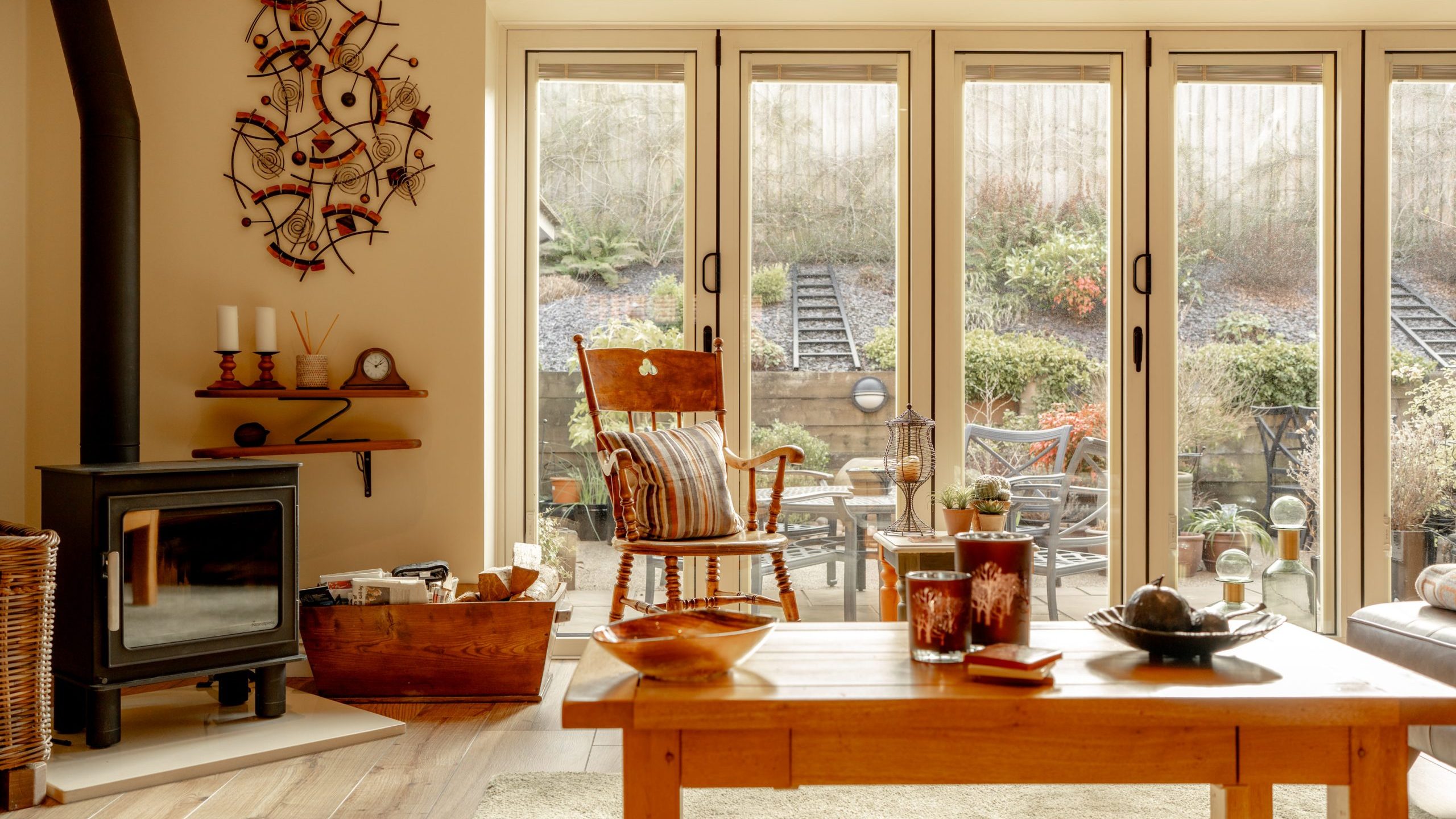 A cozy living room featuring a wooden rocking chair, a log burner with logs nearby, and a large wooden coffee table. The room opens to an outdoor patio through glass folding doors, revealing a garden with outdoor furniture and plants.