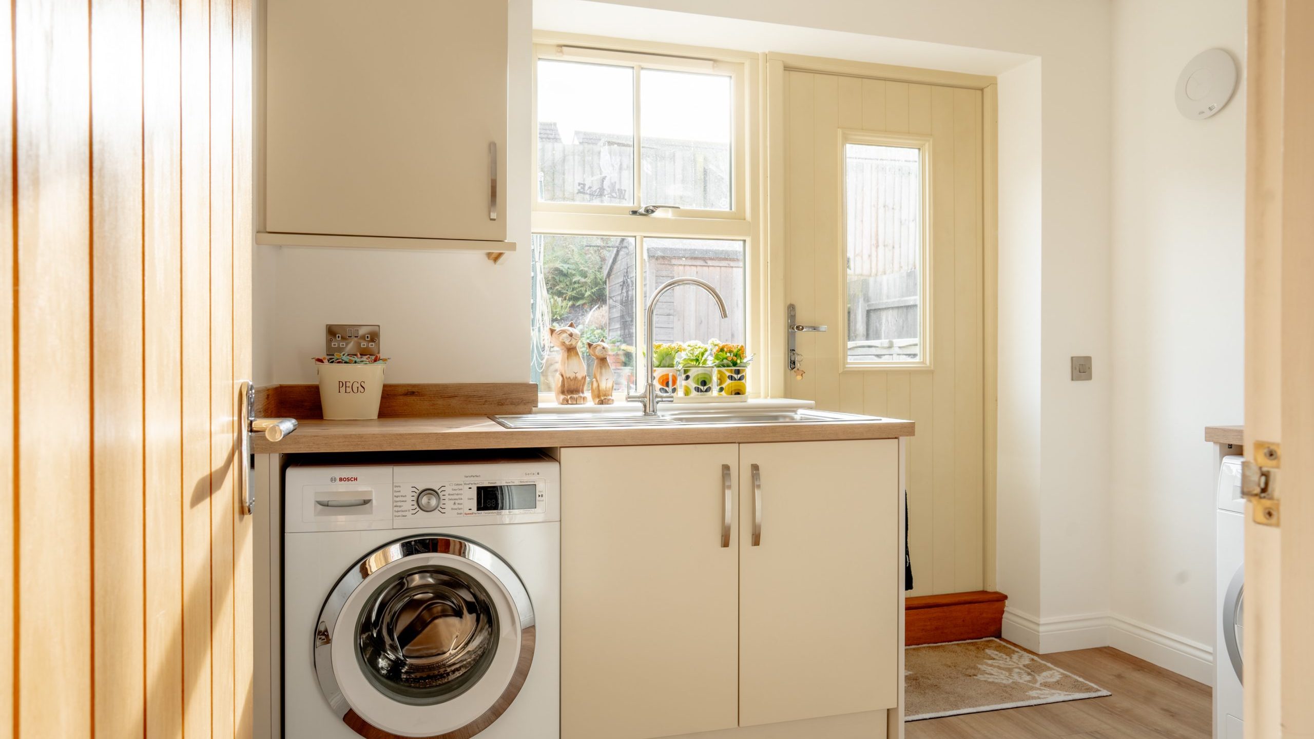 A bright laundry room with a front-loading washing machine, light wooden cabinets, and a beige countertop. A window above the counter lets in natural light, with potted plants. A door with a glass pane leads outside. Brown wooden flooring is visible.