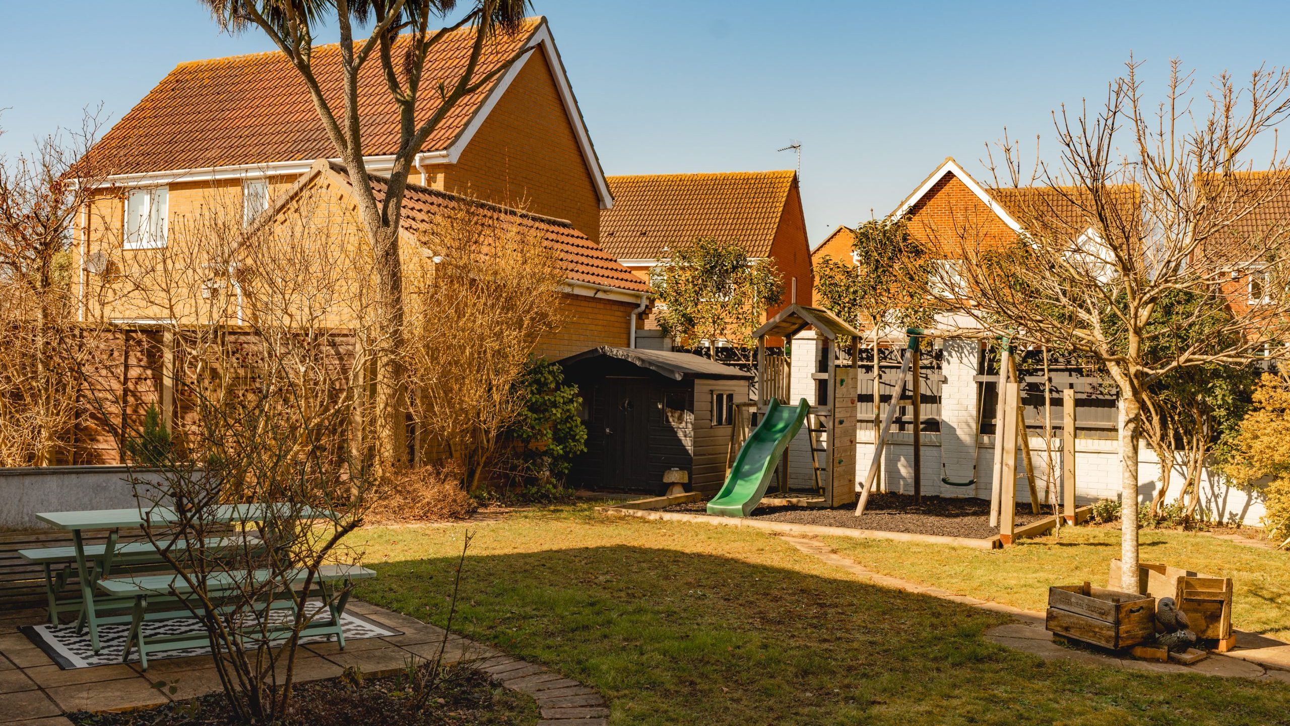 A suburban backyard with a small playground featuring a slide and climbing frame. The yard has a grassy area, a few leafless trees, and a patio with a picnic table. Several houses with red-tiled roofs are in the background under a clear blue sky.