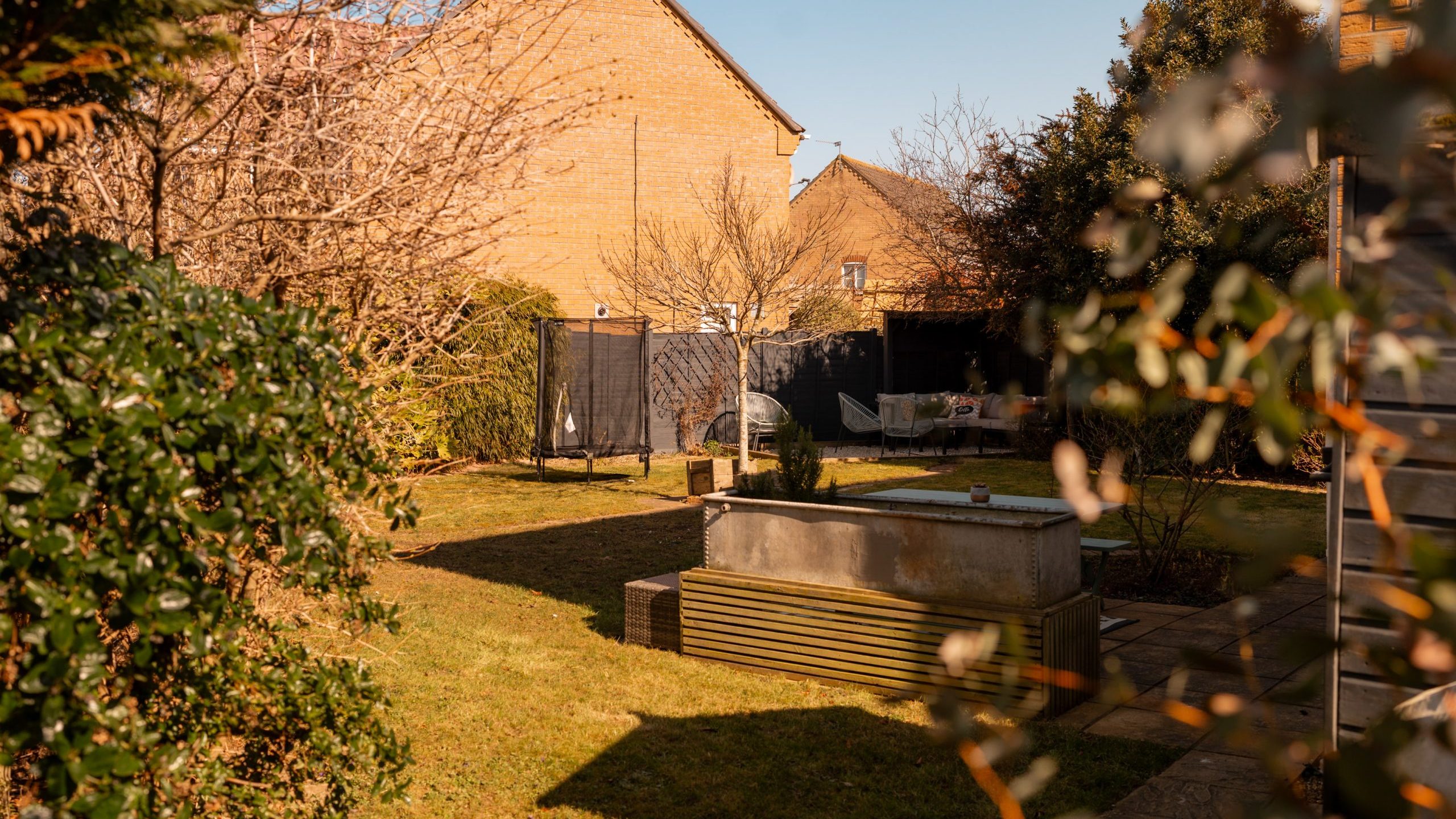 Sunny backyard with green grass, surrounded by shrubs and trees. A raised planter sits in the center, with brick houses and a garden fence in the background. The area is well-maintained and peaceful.
