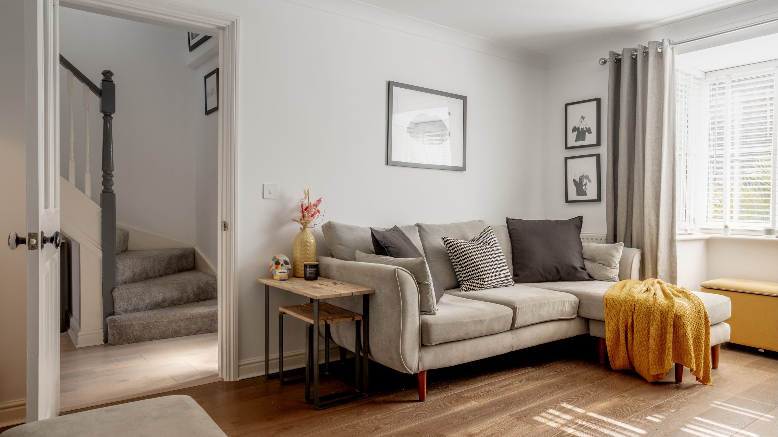 A cozy living room with a light gray sectional sofa adorned with black and white pillows and a mustard throw blanket. Wood flooring, a side table with decor, framed wall art, a window with blinds, and stairs in the background complete the scene.