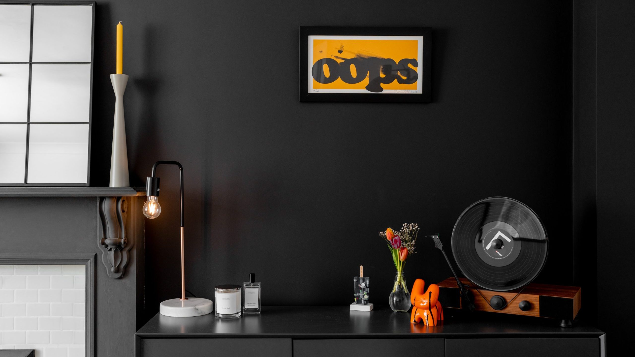 A stylish black room features a sideboard with a vintage reel-to-reel tape player, a bouquet in a glass vase, a small orange elephant figurine, and framed art titled