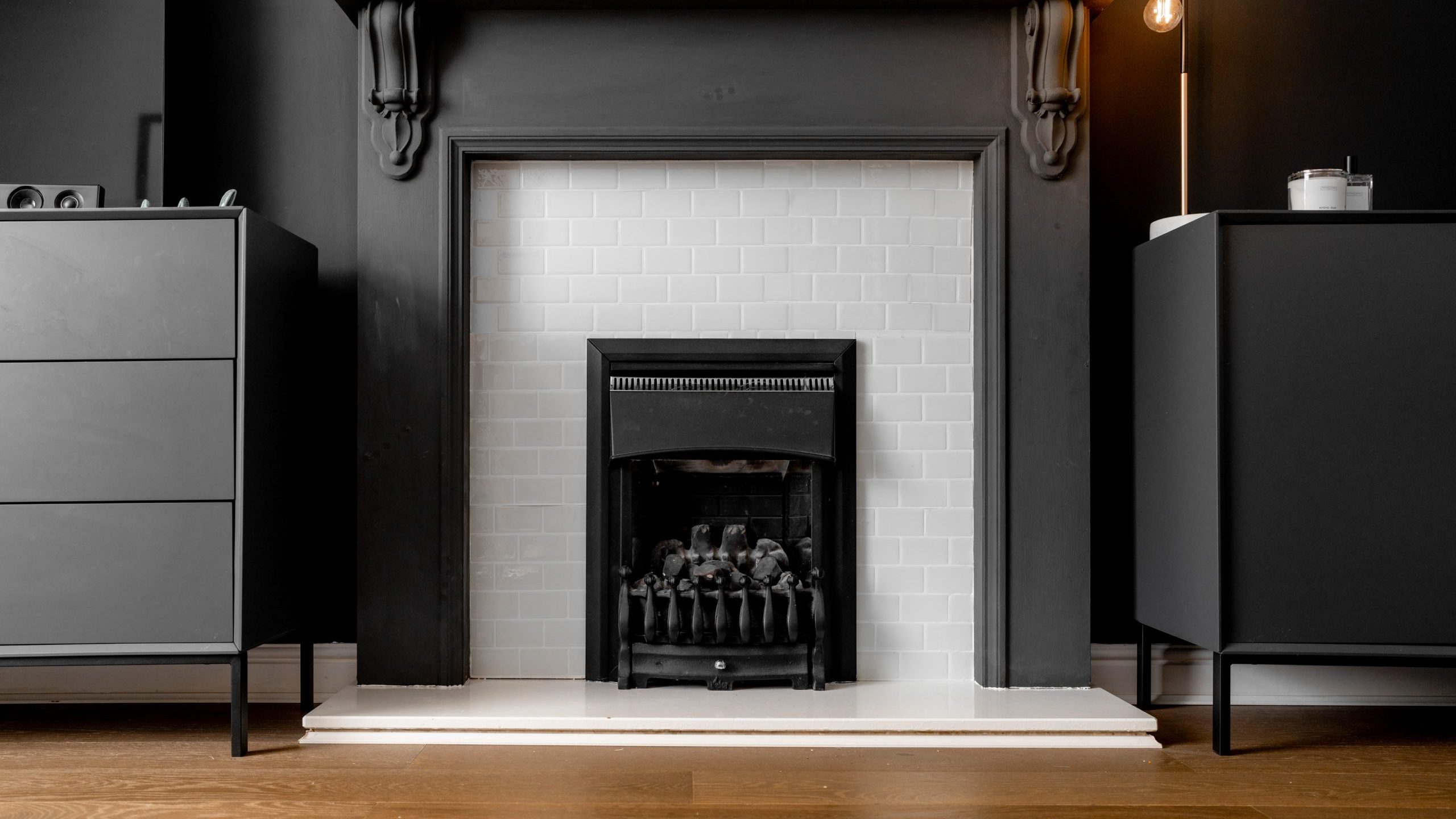 A modern living room features a dark gray fireplace with a sleek black mantel, contrasted by white subway tiles inside. On either side, minimalistic black cabinets sit against the gray walls. Warm wooden flooring adds a cozy touch.