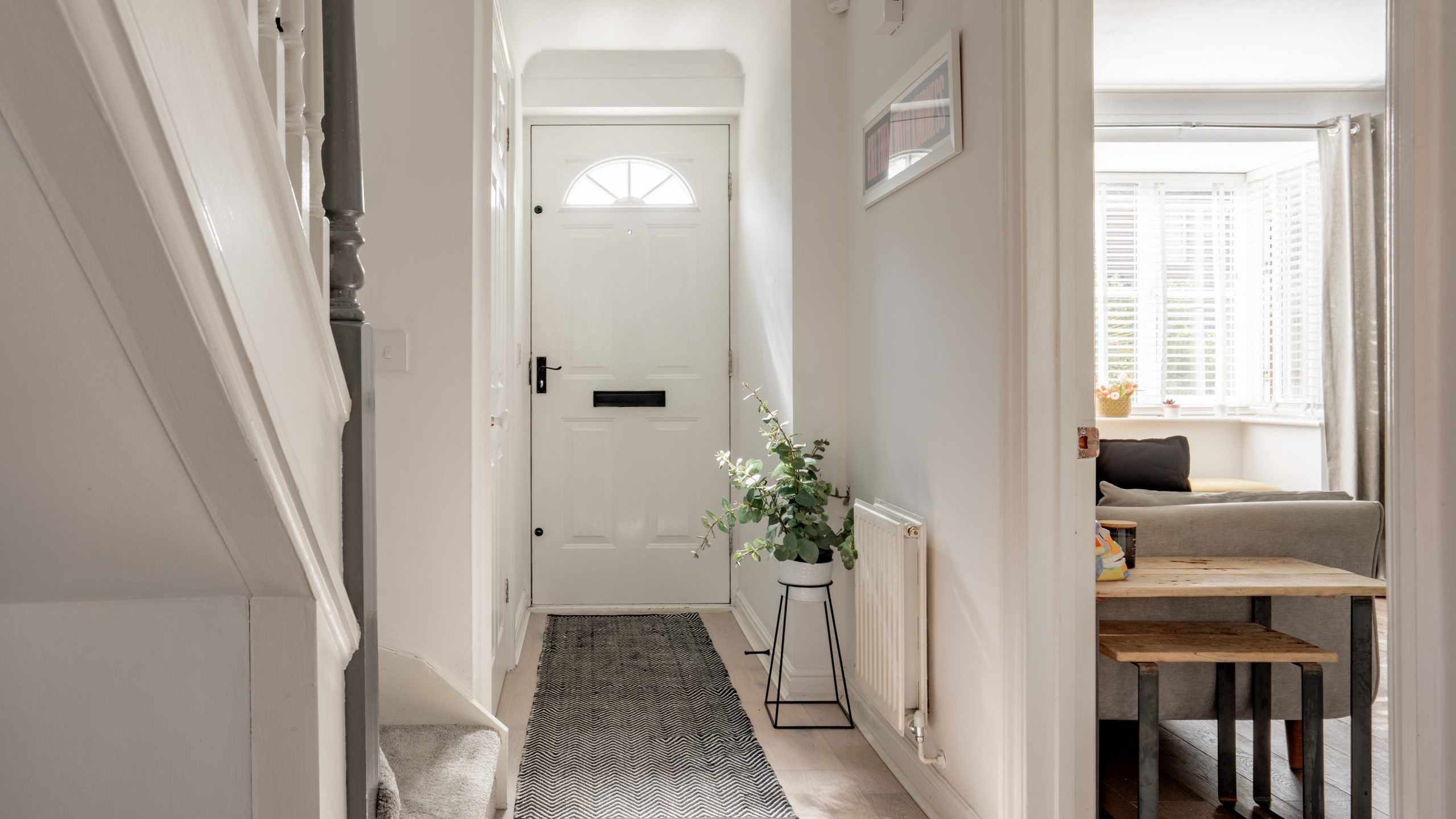 A bright hallway with a white front door and a gray doormat leads to a living room. Stairs are visible on the left, and a small plant sits on a stand. The living room has a gray sofa and wooden side tables near white shutters.