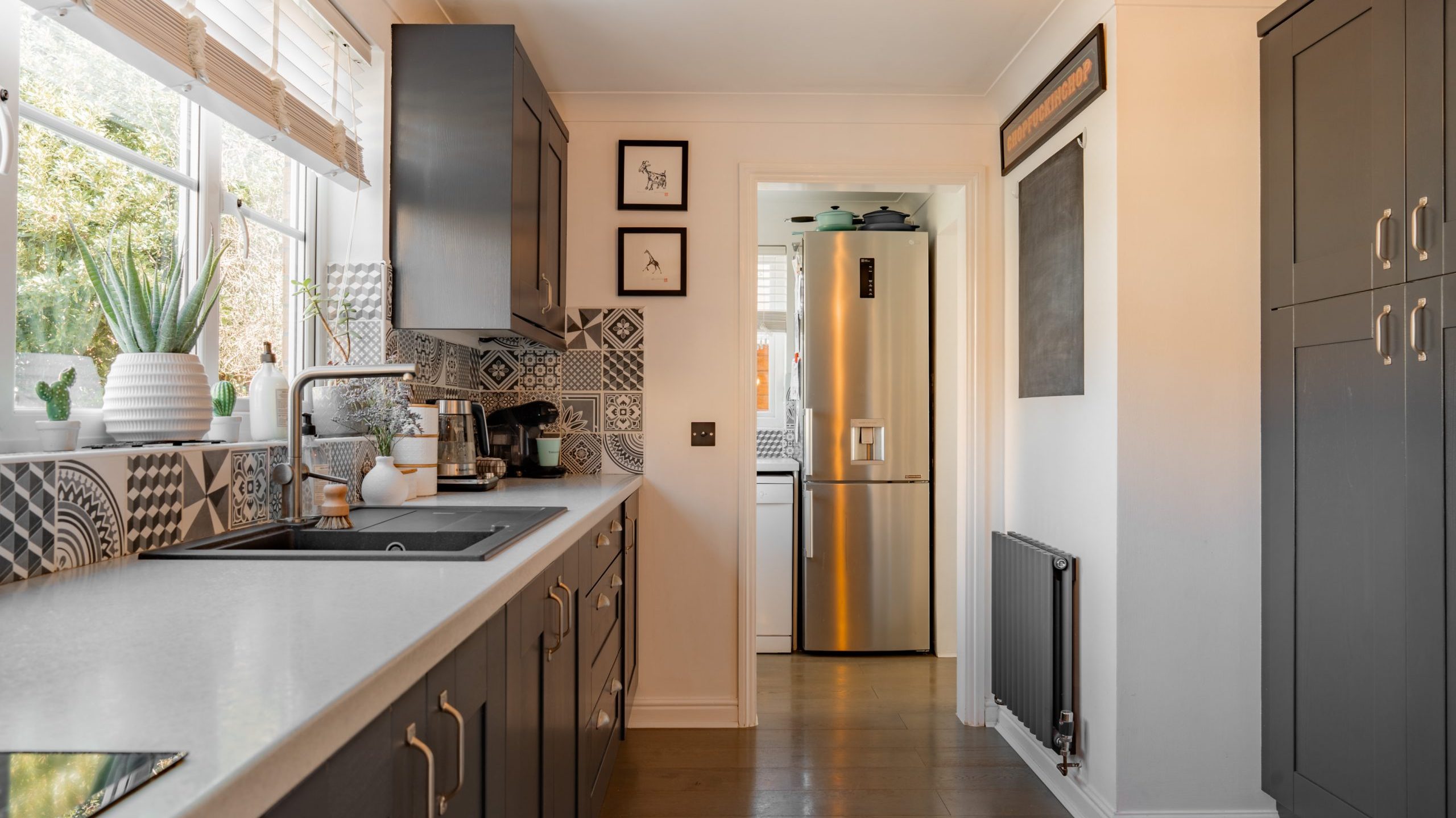 A modern, narrow kitchen with dark gray cabinets, white countertops, and a patterned backsplash. A stainless steel refrigerator stands at the far end. The room is well-lit with natural light from a window and features potted plants on the windowsill.