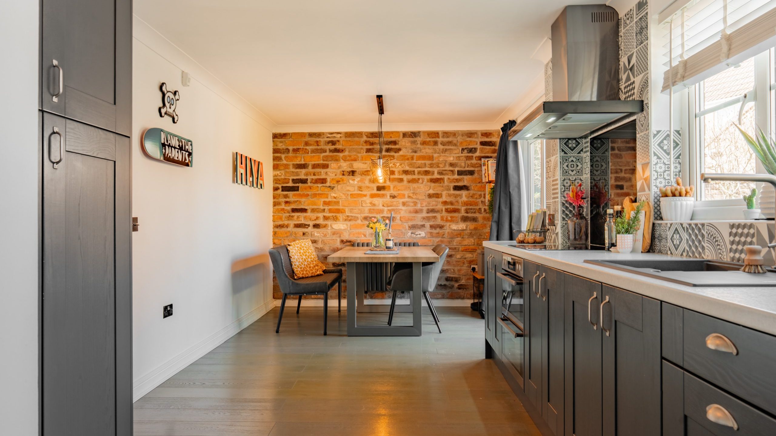 Modern kitchen with gray cabinets, tiled backsplash, and a sink on the right. Dining area features a wooden table, chairs, and a brick accent wall. Decor includes wall art and a potted plant. Large windows allow natural light to fill the space.
