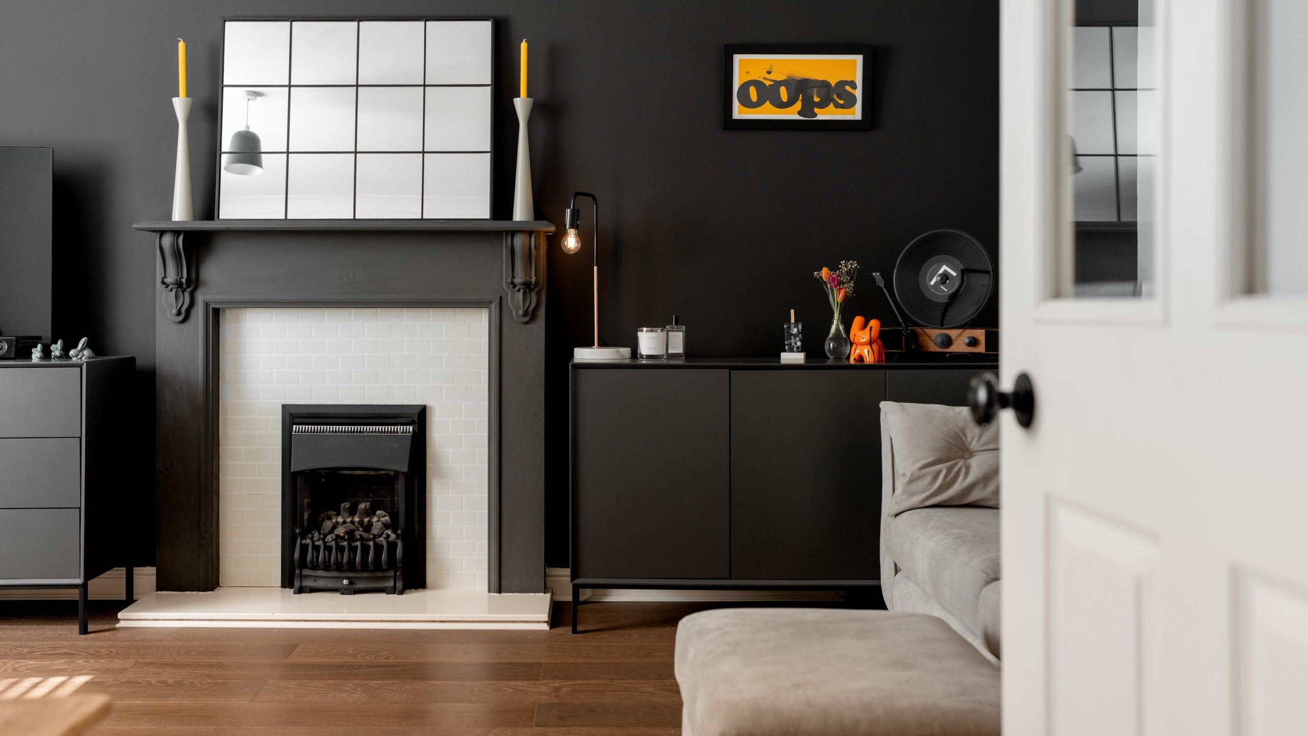 A modern living room with dark walls features a white-tiled fireplace, black console table, and minimalist decor. There's a couch, abstract wall art reading