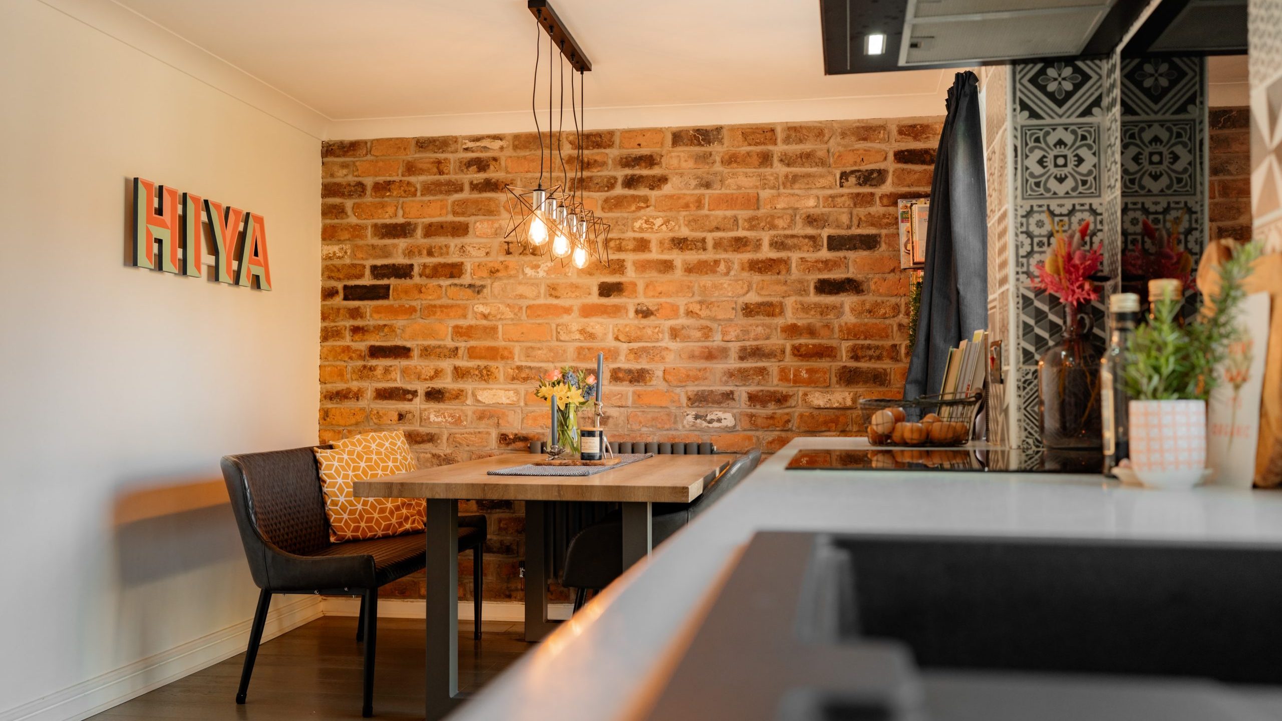 A cozy dining area with a brick accent wall. A wooden table is surrounded by chairs, with decorative lights hanging above. A sign reads
