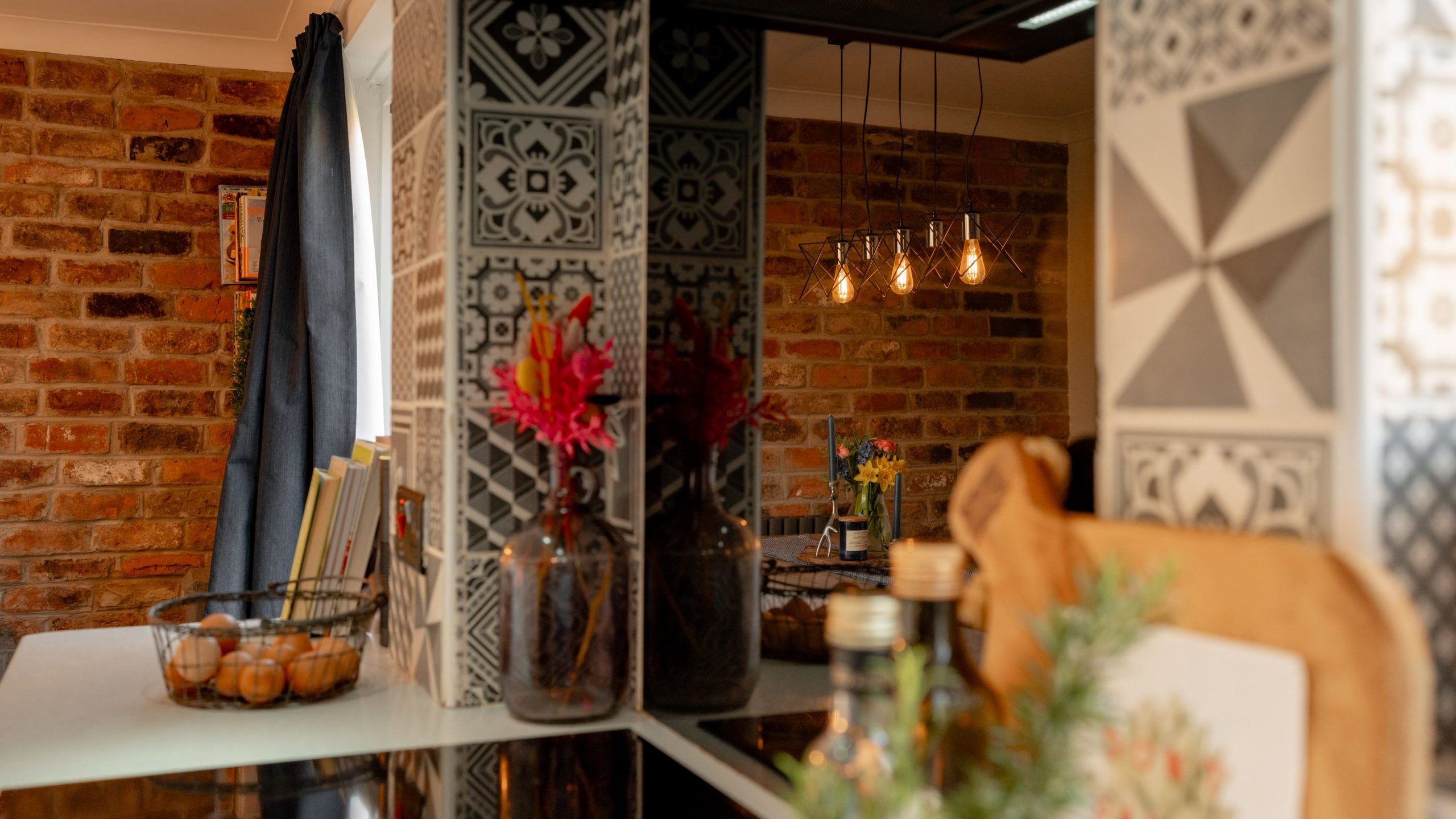 A cozy kitchen with a mix of modern and rustic design elements. The space features decorative patterned tiles, exposed brick walls, and hanging light bulbs. A vase with red flowers and kitchen items like books, jars, and a plant add warmth.