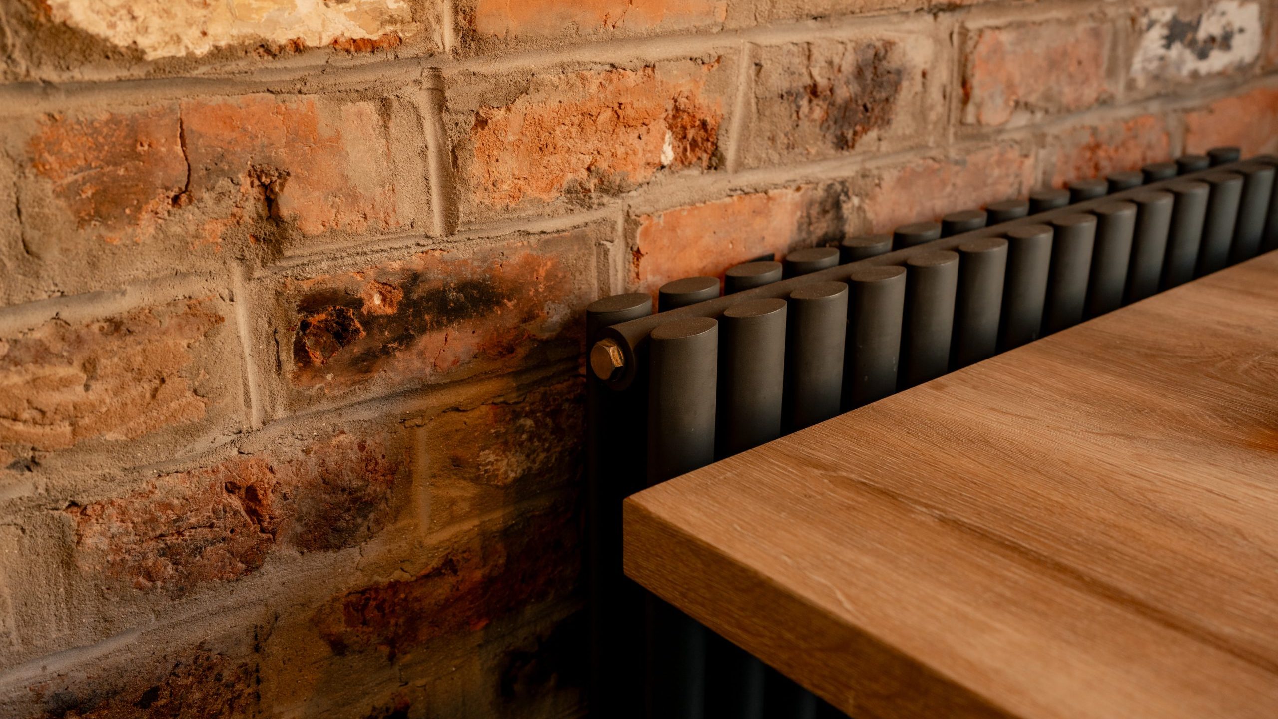 A close-up view of a dark radiator against a red brick wall, with a wooden tabletop partially visible. The bricks have a rustic, textured appearance, and the radiator has a sleek, vertical design.