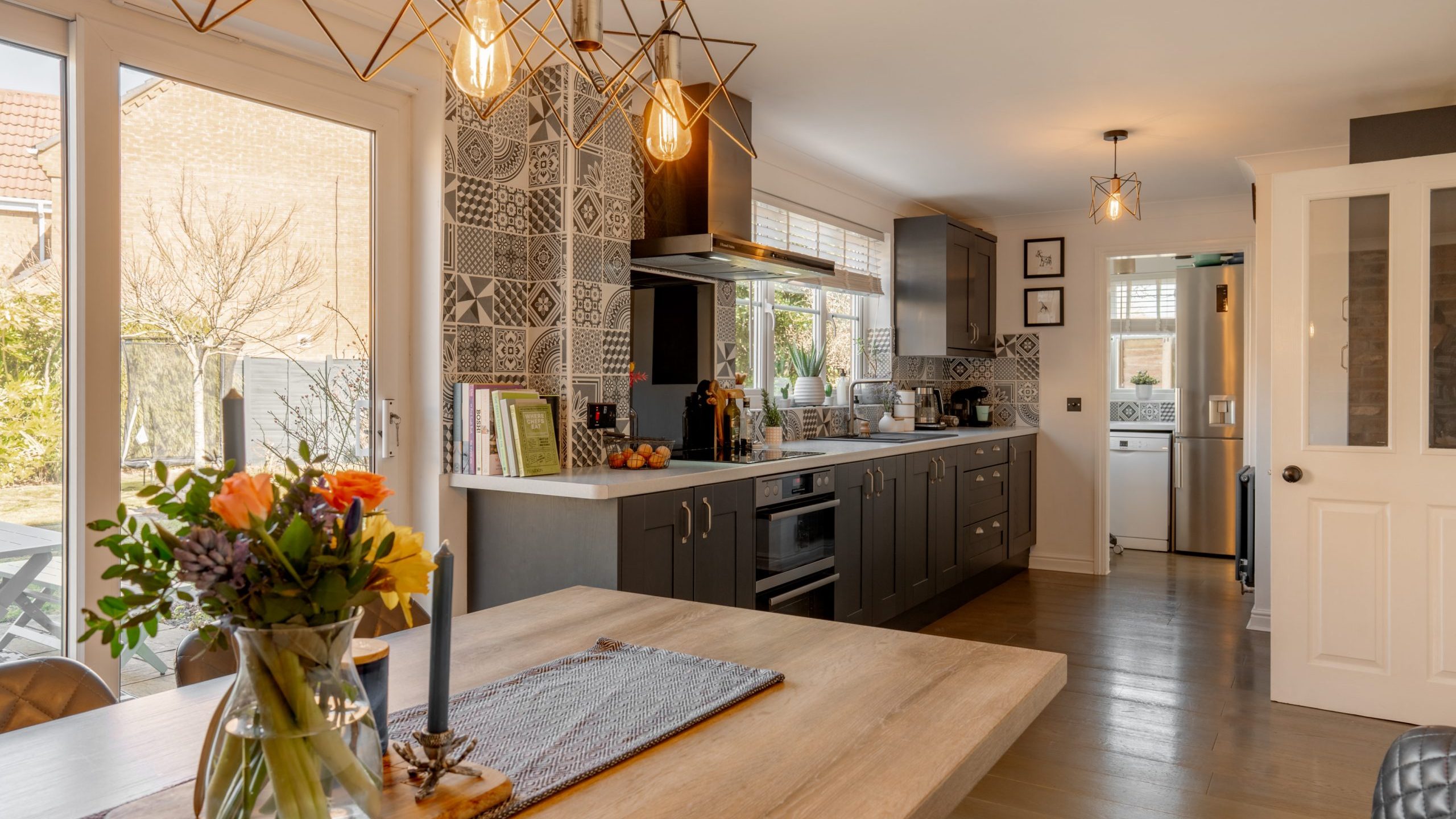 A modern kitchen with patterned tiles and dark cabinets. The room is well-lit with unique pendant lights above the wooden dining table, which has a vase of flowers. A window reveals a garden outside, and a glimpse of a laundry area is visible through a doorway.