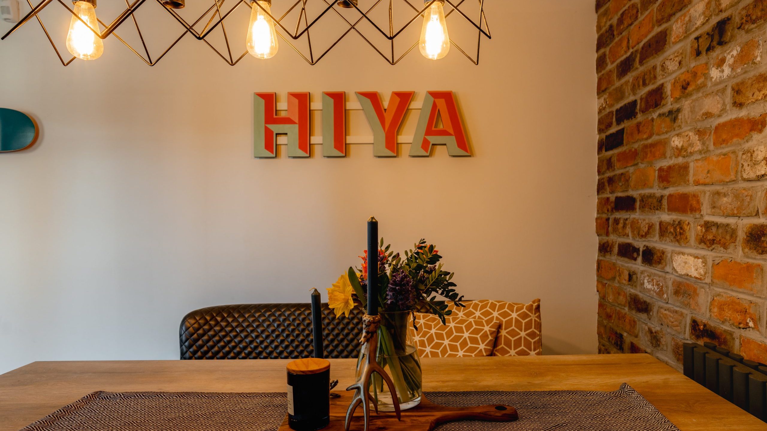 Dining area with a wooden table set with a black candle and flowers in a vase. Exposed brick wall on the right. Decorative lights hang above, and the word
