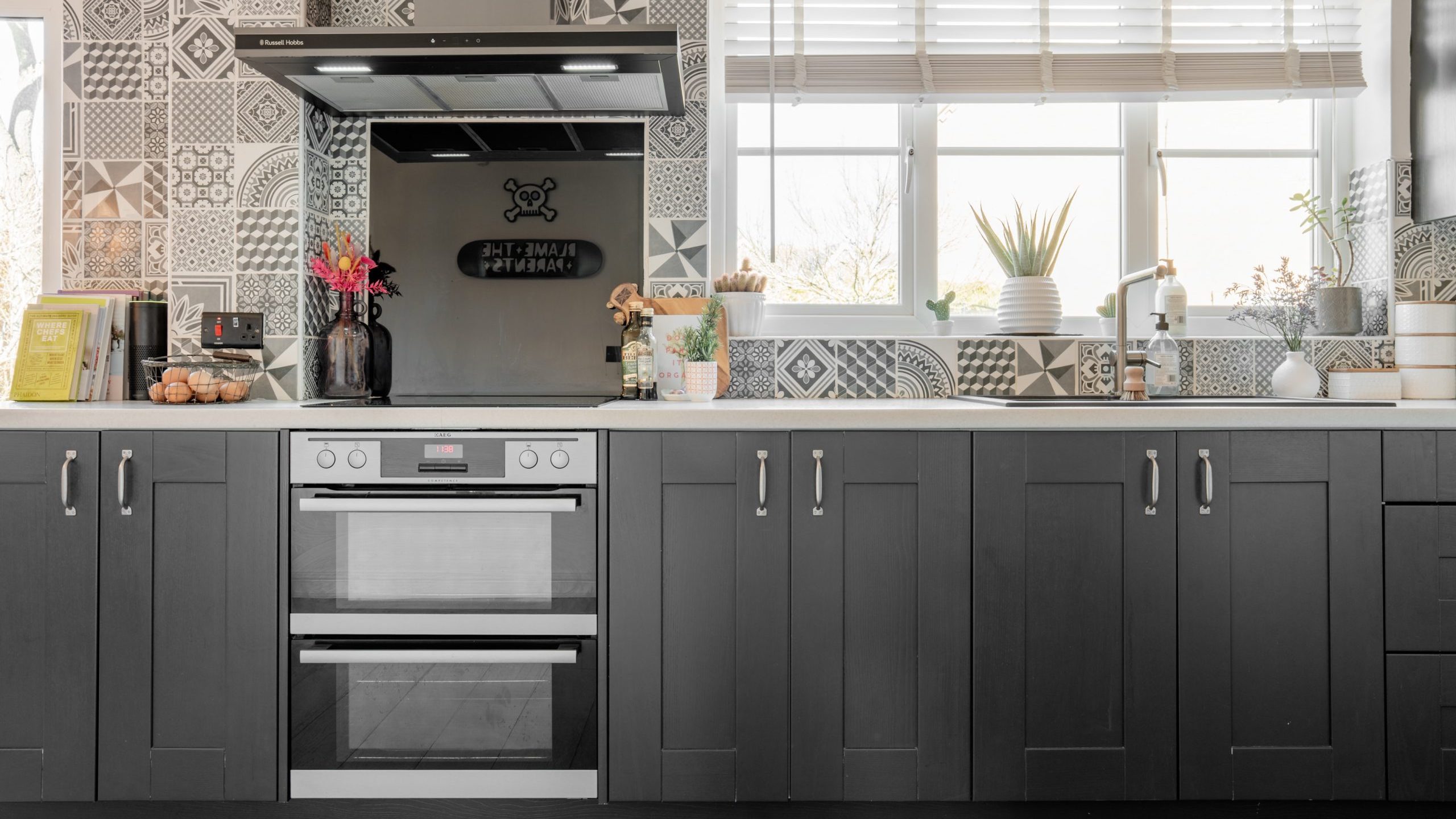 Modern kitchen with dark cabinets, a double oven, and a patterned tile backsplash. A vase with pink flowers, a potted plant, and kitchen items are on the counter. Bright window with blinds and planters.