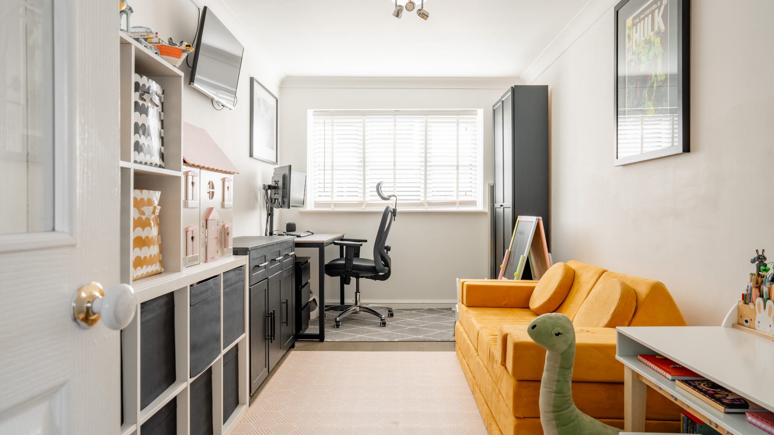 Bright room with a yellow sofa, dinosaur plush toy, and wall art. A desk with a computer and chair sits by a window. Shelving units display books and storage boxes. Window blinds allow natural light. Modern, cozy workspace and play area.