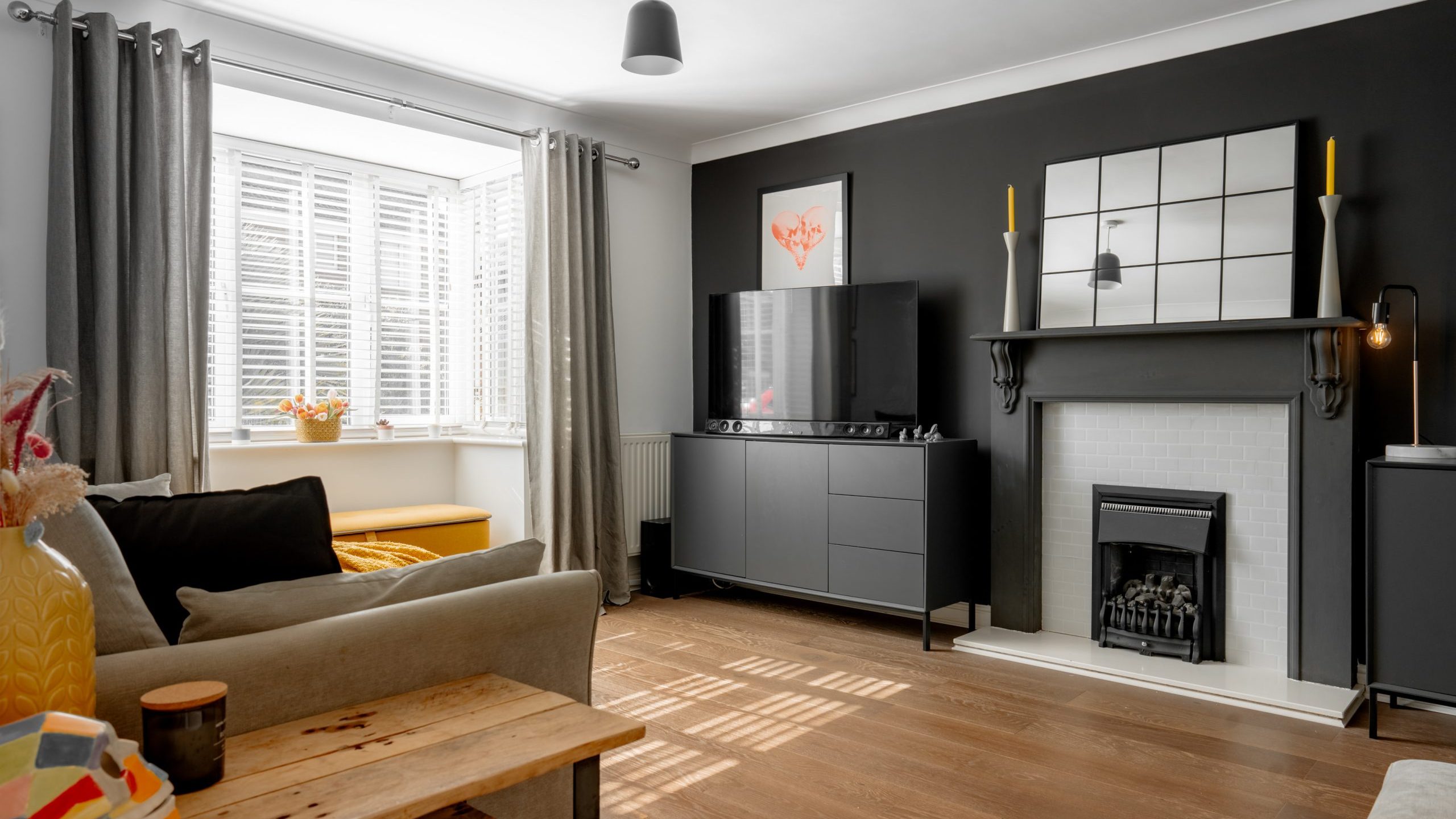 Modern living room with wooden flooring, gray sofa, and a wooden coffee table. A large TV stands on a gray console against a dark accent wall. There’s a fireplace with a decorative mirror above, and a bay window with curtains and blinds.