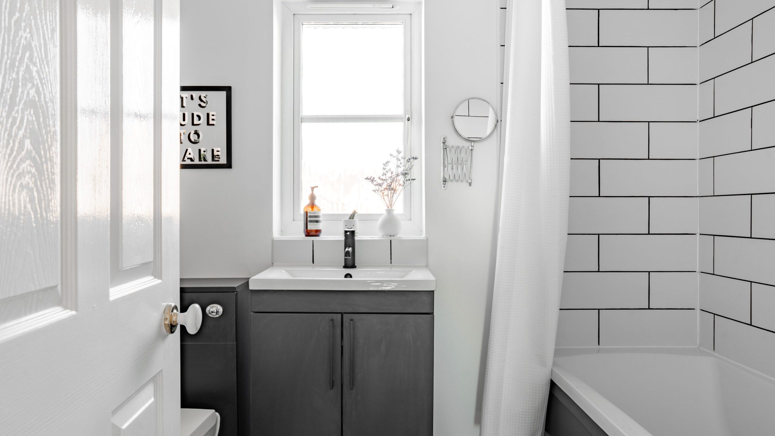 Minimalist bathroom with white subway tiles and a white shower curtain. A sleek gray vanity with a mirror and small vase of flowers sits beneath a window. A framed sign hangs on the wall. The room has a clean, modern look.