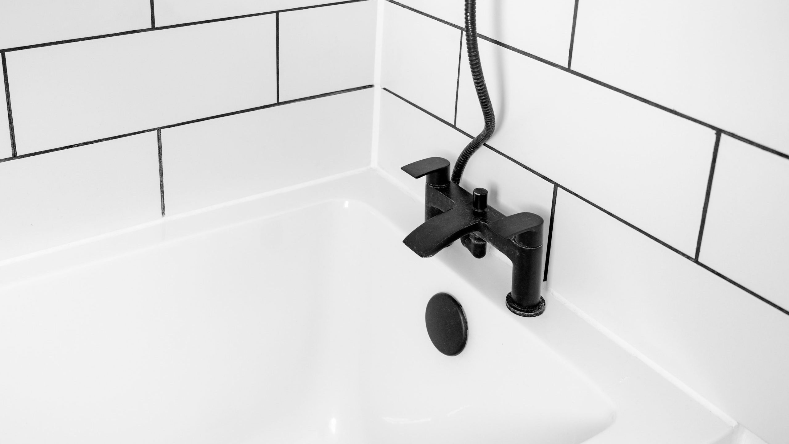 A modern white bathtub with black fixtures, including a faucet and a shower head. The surrounding walls are tiled with large white tiles divided by black grout, creating a sleek, minimalist design.