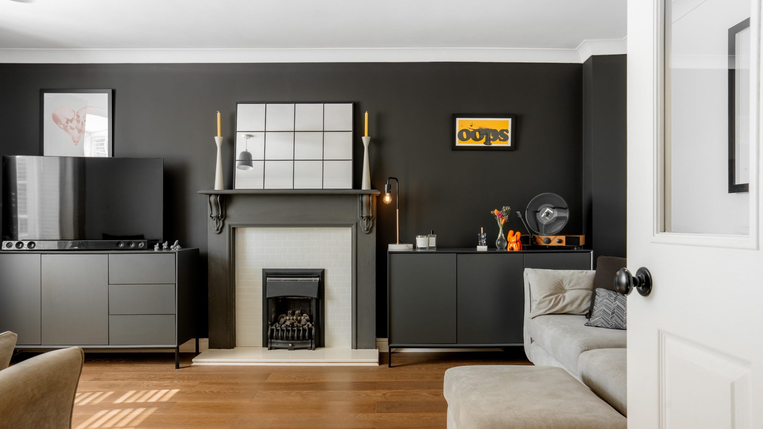 A modern living room with dark walls features a fireplace, mirror, and decorative items on the mantel. A TV and cabinets are on the left, and a beige sofa is on the right. A white door is partially open, revealing a wooden floor.
