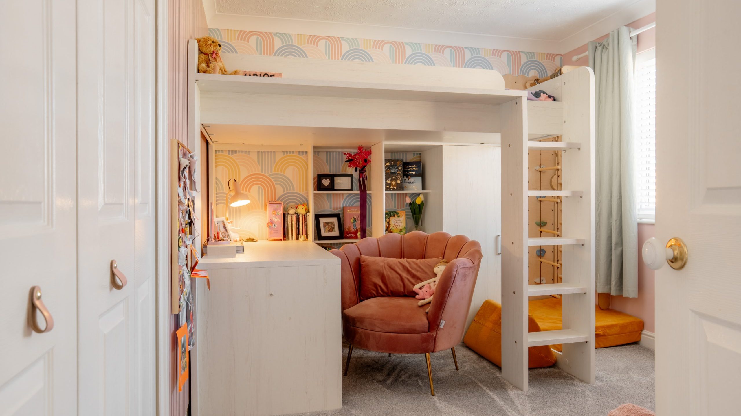 A cozy room with a loft bed and a colorful geometric wallpaper. Below the bed is a plush pink chair and a white desk with books and plants. An orange beanbag lies on the carpeted floor near a window with light curtains.