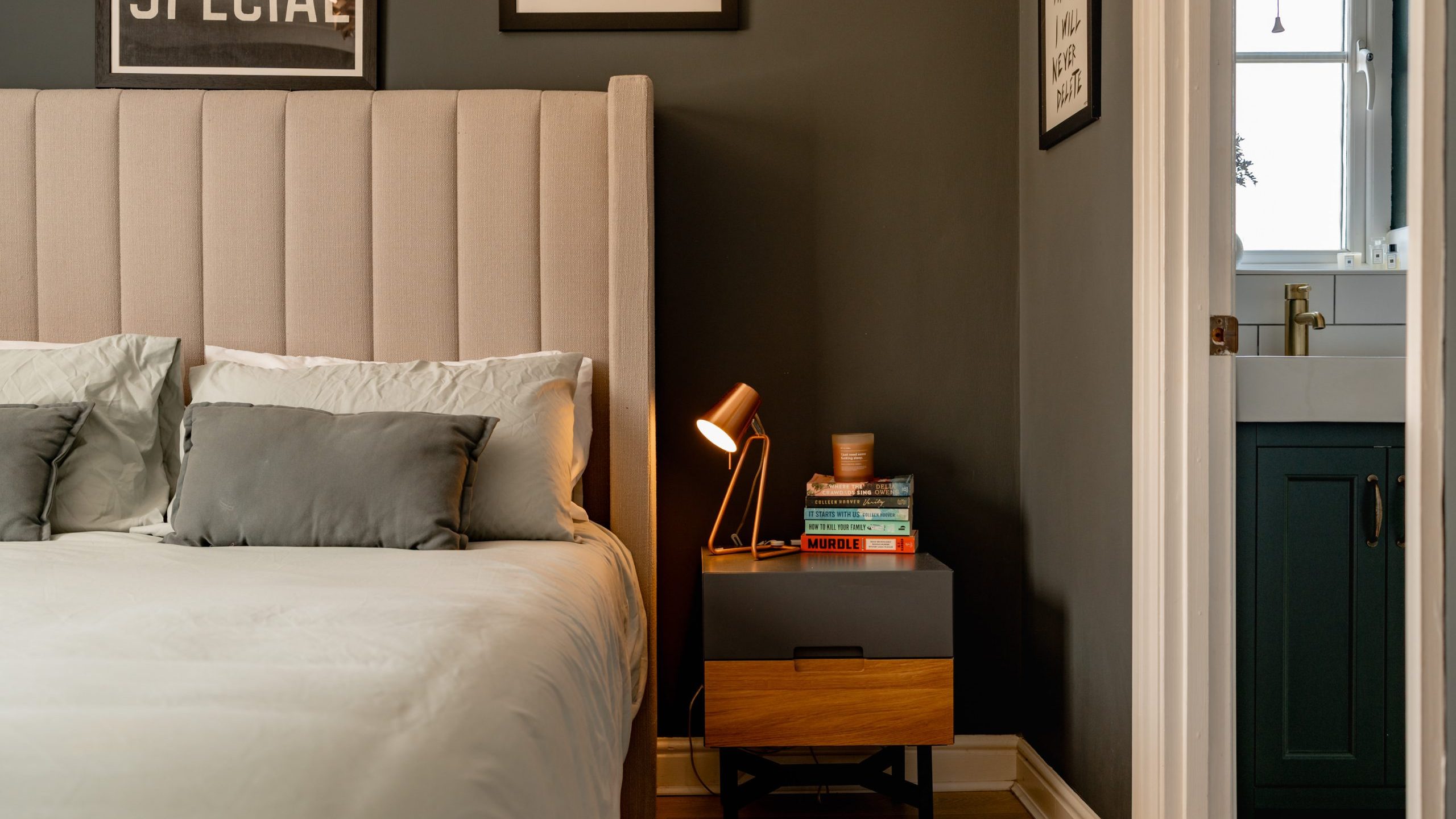 A cozy bedroom with a beige upholstered headboard, light gray bedding, and a black and wood nightstand. A small lamp illuminates the nightstand, which holds books and a candle. The room has dark gray walls and leads to a bathroom with a dark vanity.