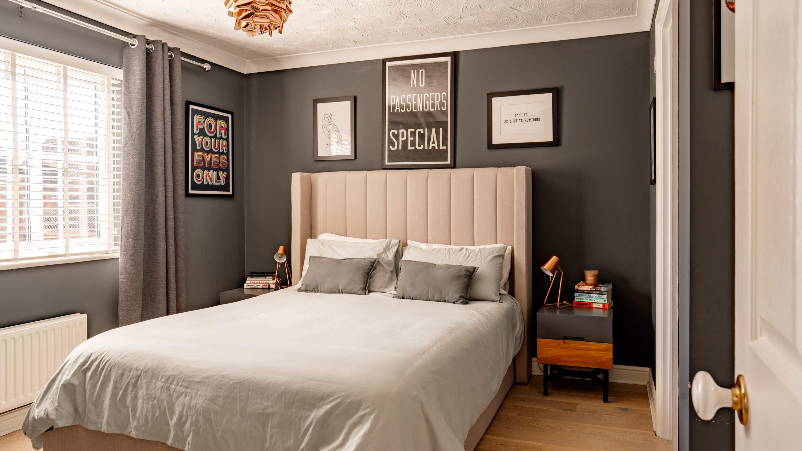 A cozy bedroom with a beige upholstered bed, light gray bedding, and wooden nightstands. The walls are dark with various framed artworks. A window on the left lets in natural light through white blinds. A wooden light fixture hangs from the ceiling.