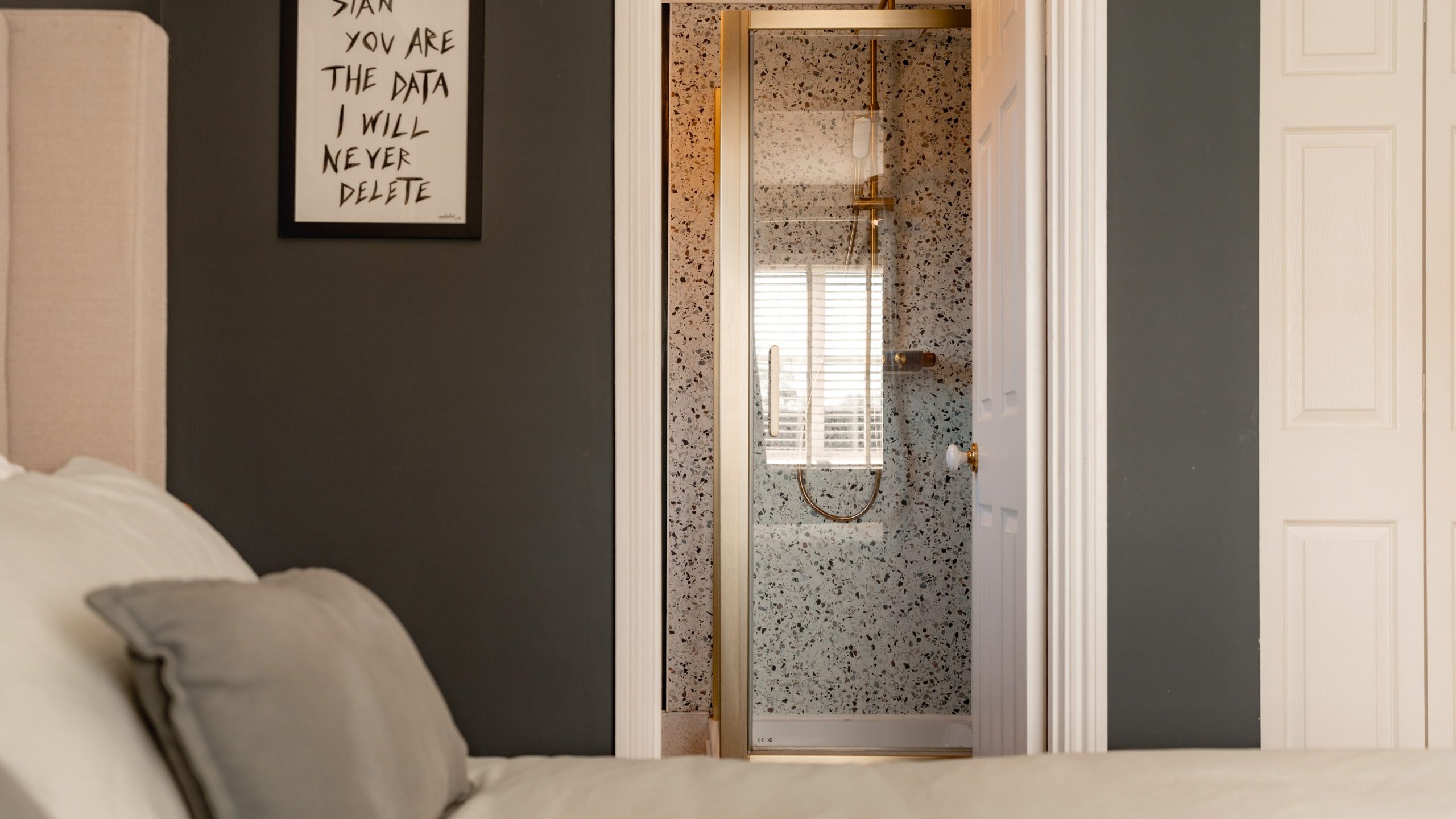 A cozy bedroom with a beige bedspread and gray accent pillows. A framed sign on the dark gray wall reads,