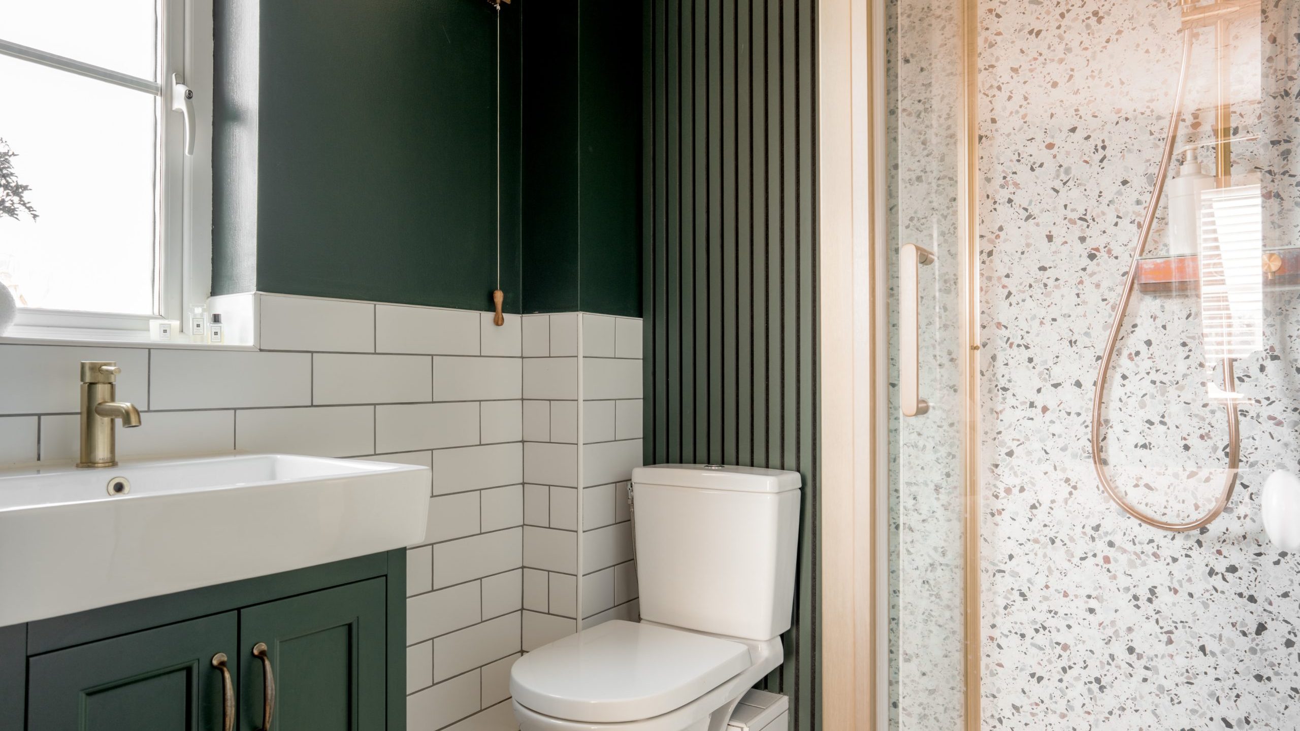 A modern bathroom with green walls and white subway tiles. It features a white toilet, a sink with brass faucet and green cabinet, and a shower with a floral-patterned glass door. A small window lets in natural light.