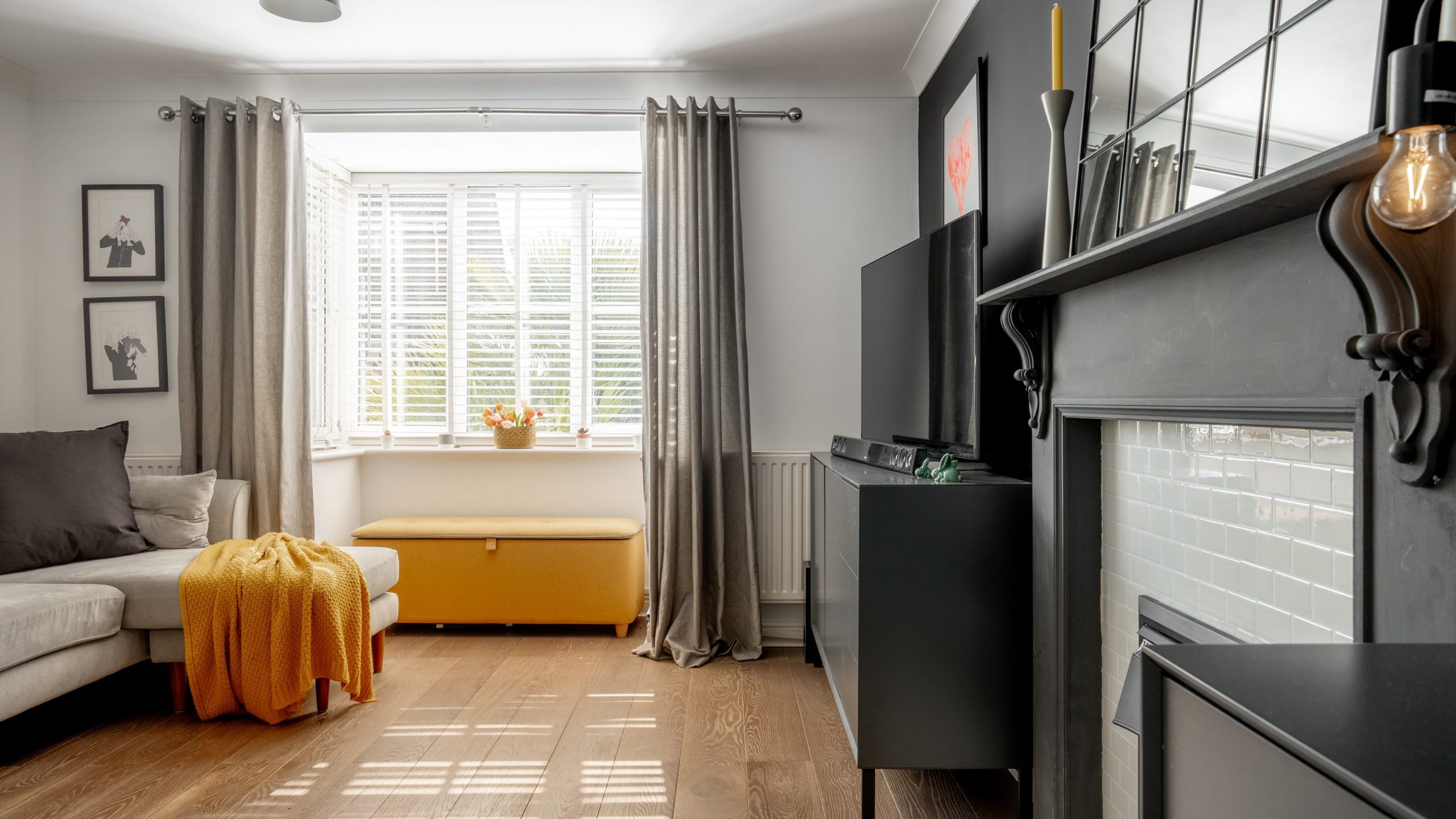 A modern living room with a gray sectional sofa, orange blanket, and a yellow bench below a window with blinds. Dark gray accents include curtains, a wall, and a cabinet. A fireplace and TV are on the right. Artworks adorn the walls.
