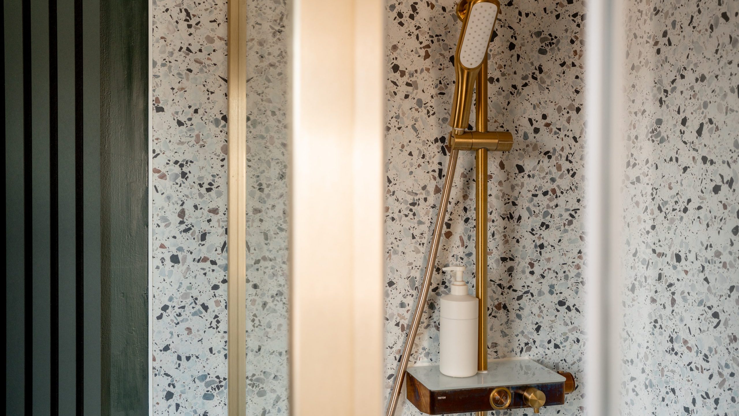 A modern shower with a terrazzo wall features a sleek gold showerhead and a matching rail. A small shelf holds a bottle, nestled in the elegant, minimalist bathroom design.