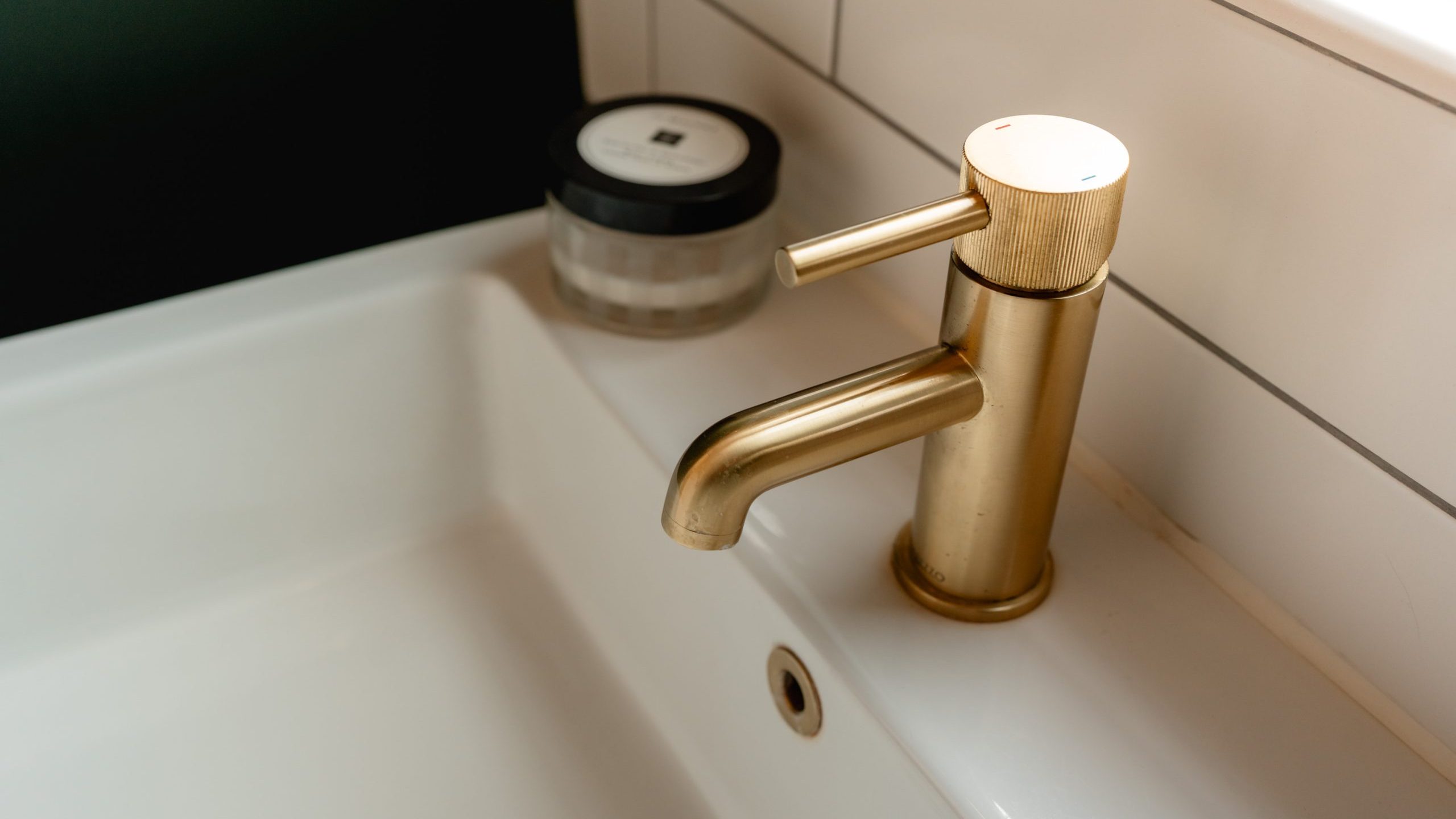 A white bathroom sink with a sleek gold faucet. A black and white container is placed on the edge of the sink. The backdrop features white tiles and a black wall.