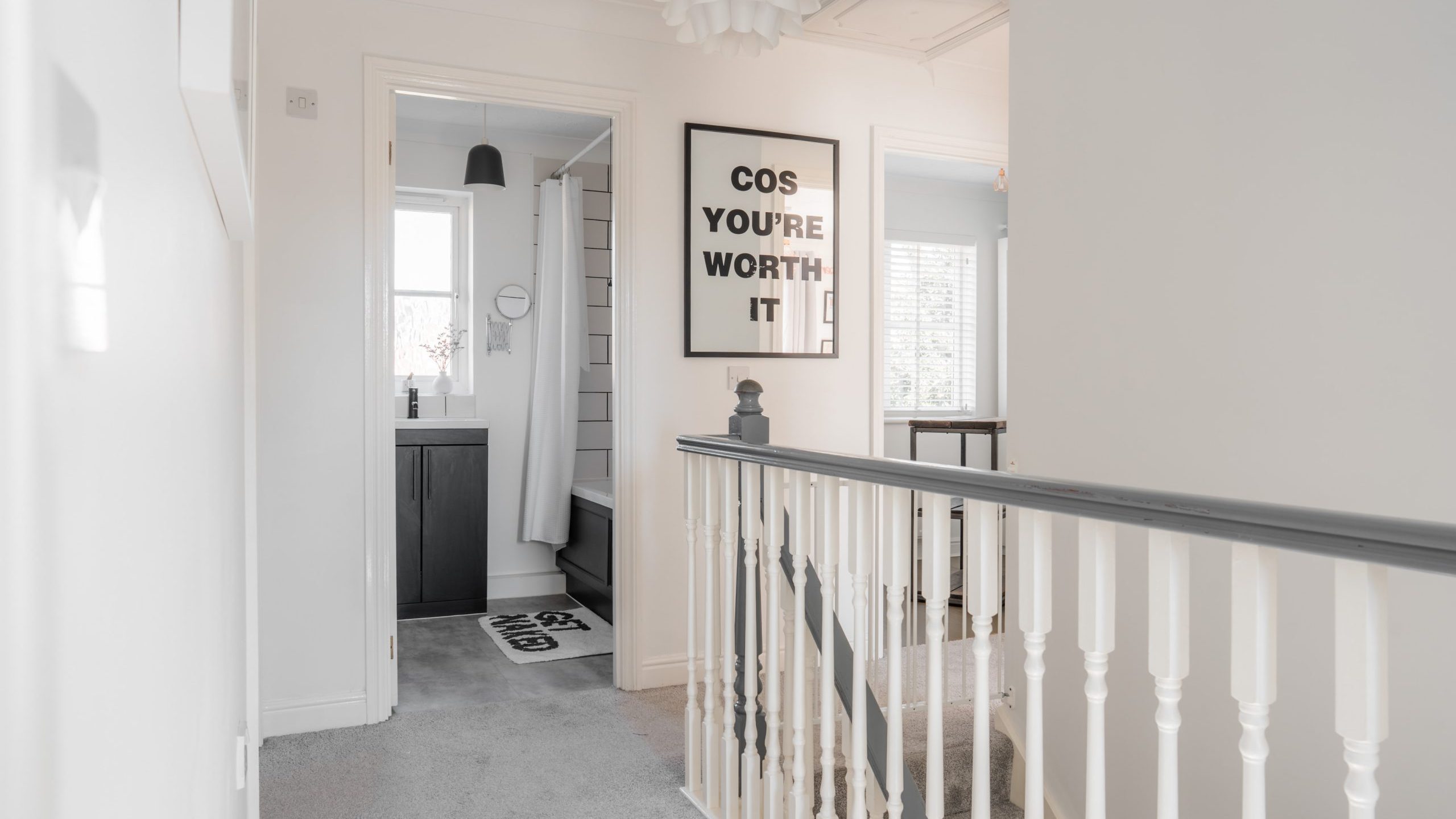 Bright hallway with white walls and carpeted floor. A staircase with a white and dark railing is on the right. Two framed quotes hang on the walls, and a bathroom with black accents is visible through an open door. A pendant light hangs from the ceiling.