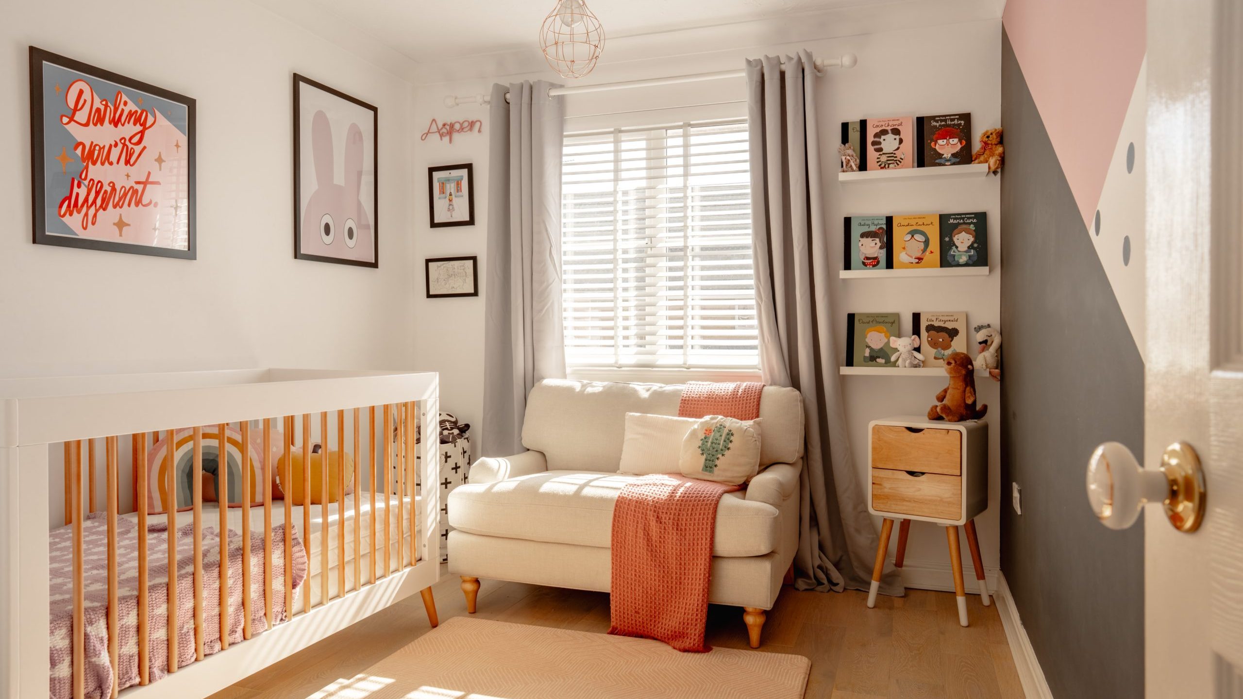 A cozy nursery with a crib next to an armchair adorned with a pink blanket. Sunlight streams through slatted blinds. Shelves hold colorful books and toys. Walls are decorated with bunny art and inspirational quotes. The room has a pink, black, and white theme.