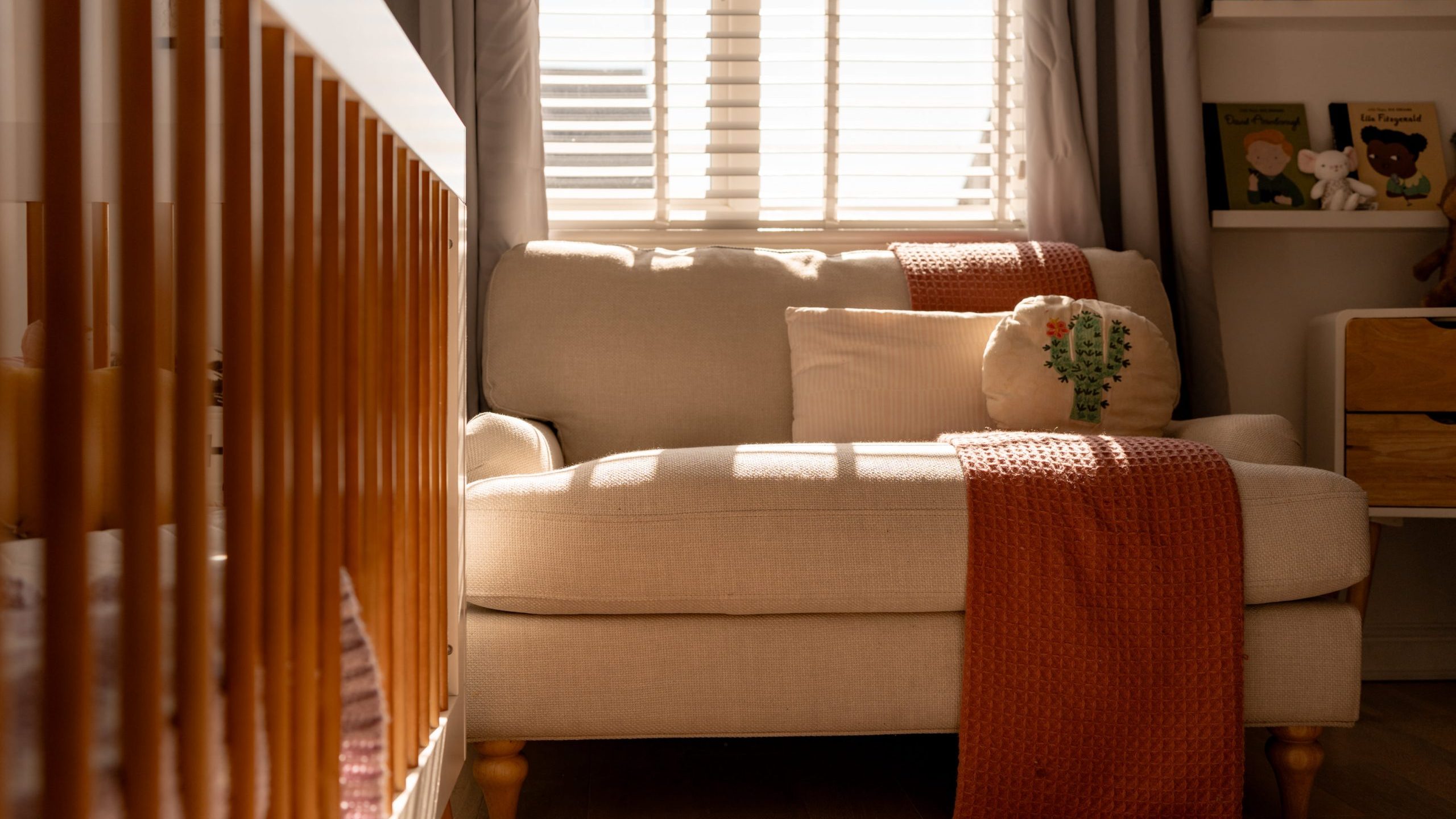 A cozy room with a beige sofa adorned with a red blanket and a cute pillow. Sunlight streams through the window, casting warm shadows. A wooden crib is in the foreground, and books and toys are on shelves in the background.