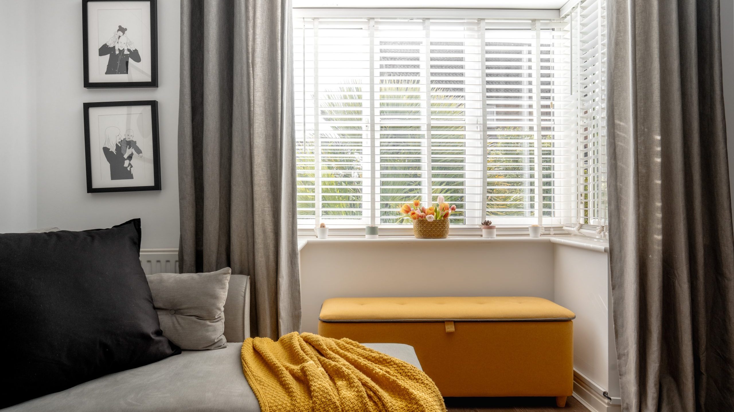 A cozy living room corner features a gray couch with a black pillow and a mustard-yellow blanket. Next to it is a yellow storage bench under a large window with blinds. Two black and white framed artworks hang on the white wall.