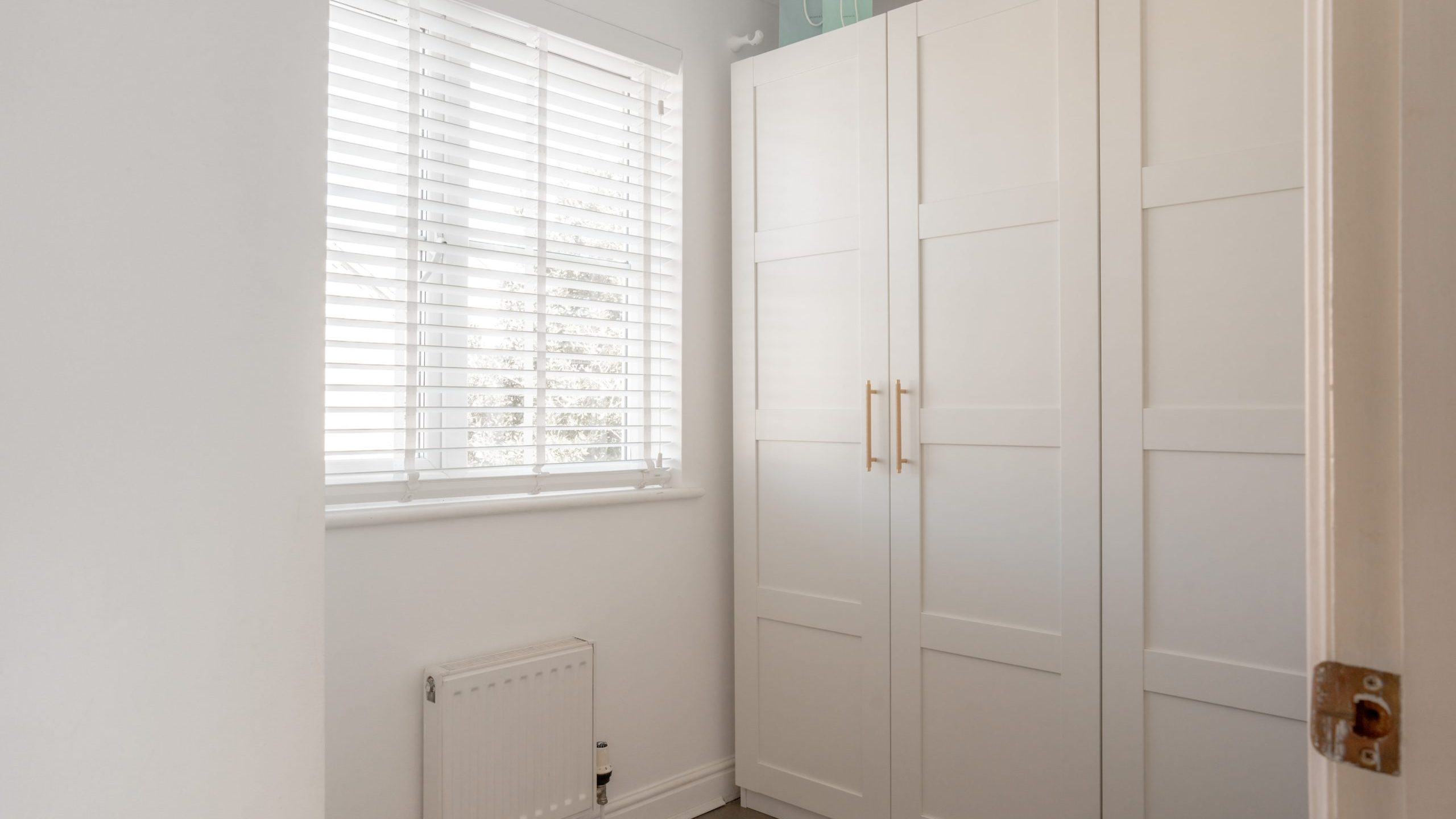 A small, bright room with white walls and a window with white blinds. There's a white wardrobe with brass handles on the right and a small radiator below the window on the left. The floor appears to be gray.