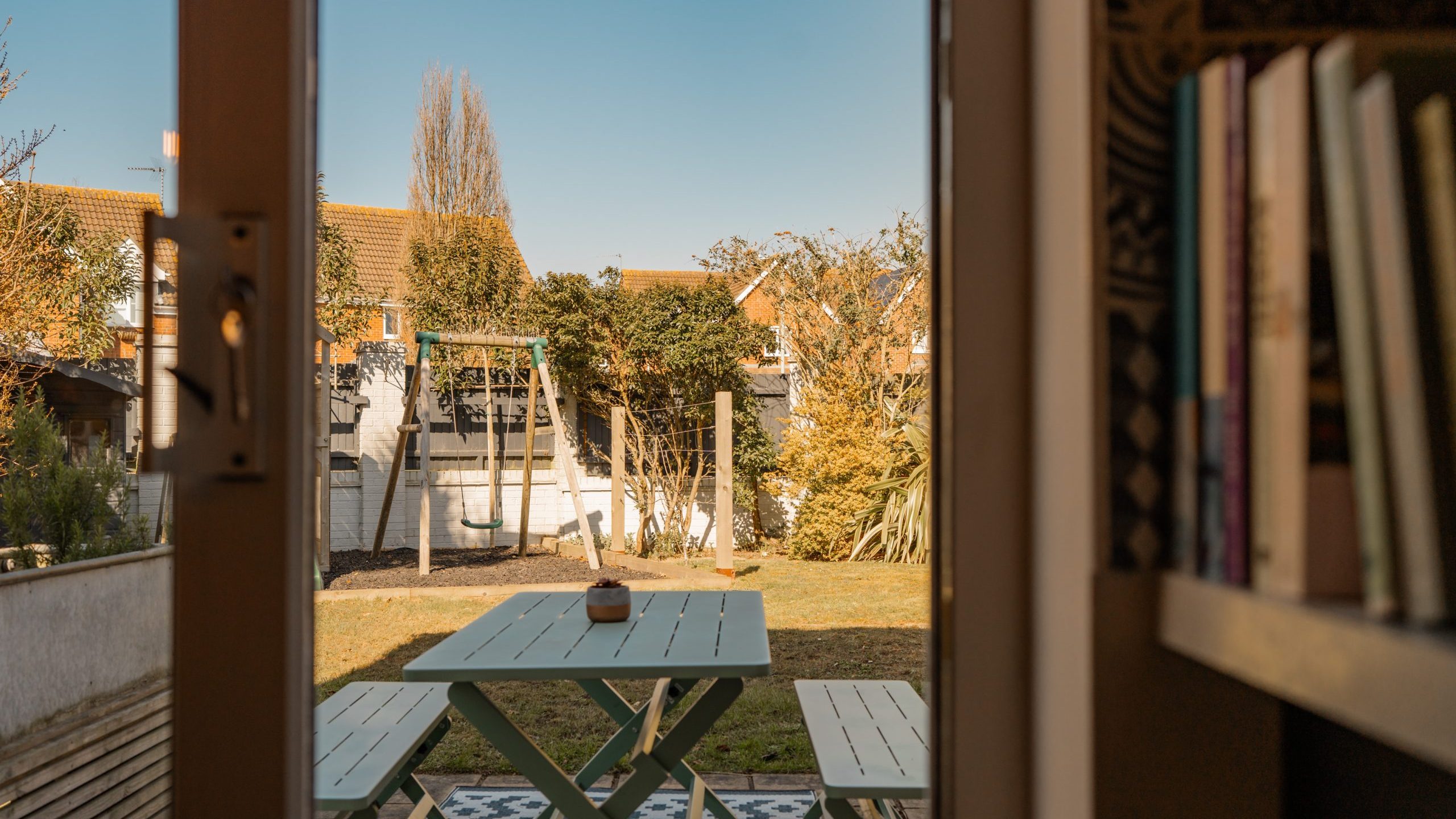 View through an open door showing a backyard with a green picnic table on a patterned patio. A swing set is visible against a backdrop of trees and a clear sky. A bookshelf is partially visible on the right.