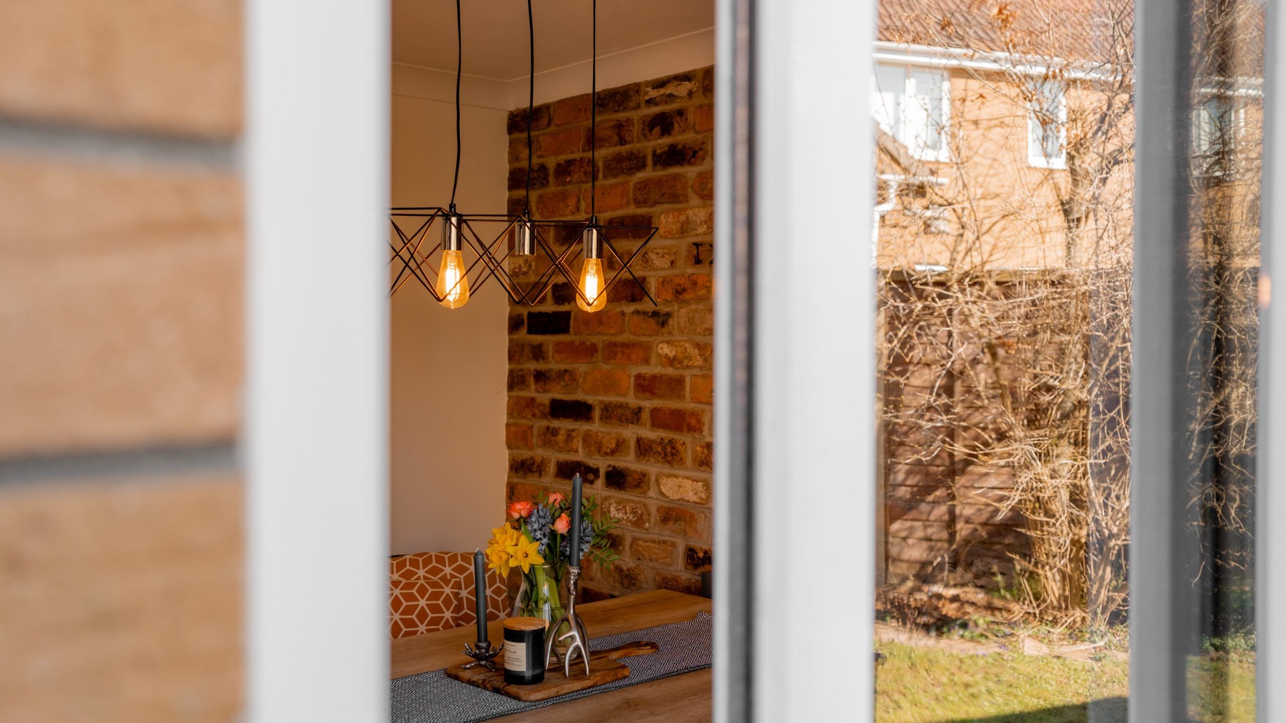 A view through a window reveals an indoor dining area with a wooden table, chairs, and a vase of flowers. Warm pendant lights hang above, illuminating a brick accent wall. Outside, a grassy yard and neighboring building are visible.