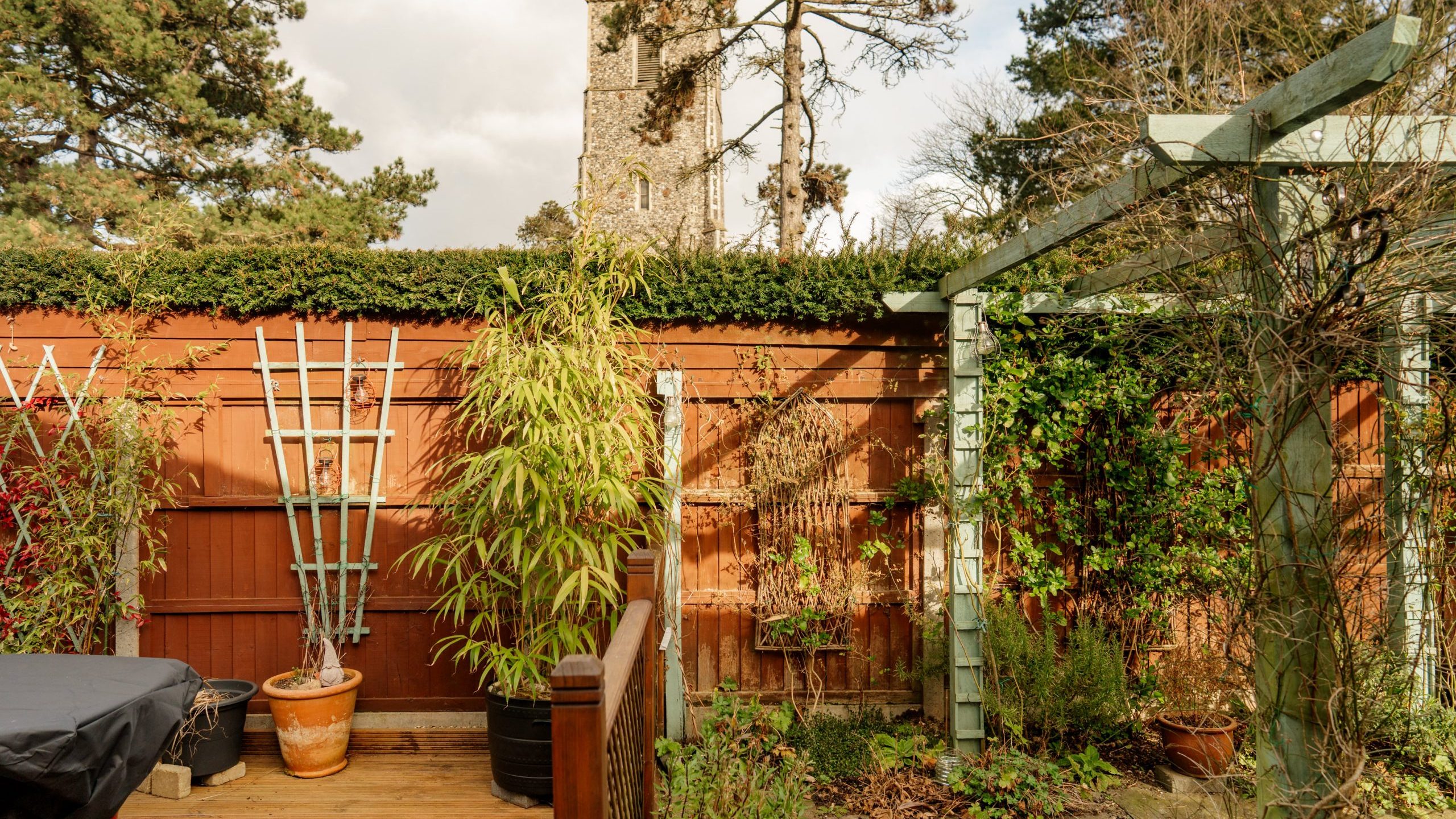 A lush garden with potted plants and a trellis sits in the foreground, while a stone structure, possibly a church tower, is visible over a wooden fence. Tall trees surround the scene, and the sky is partly cloudy.